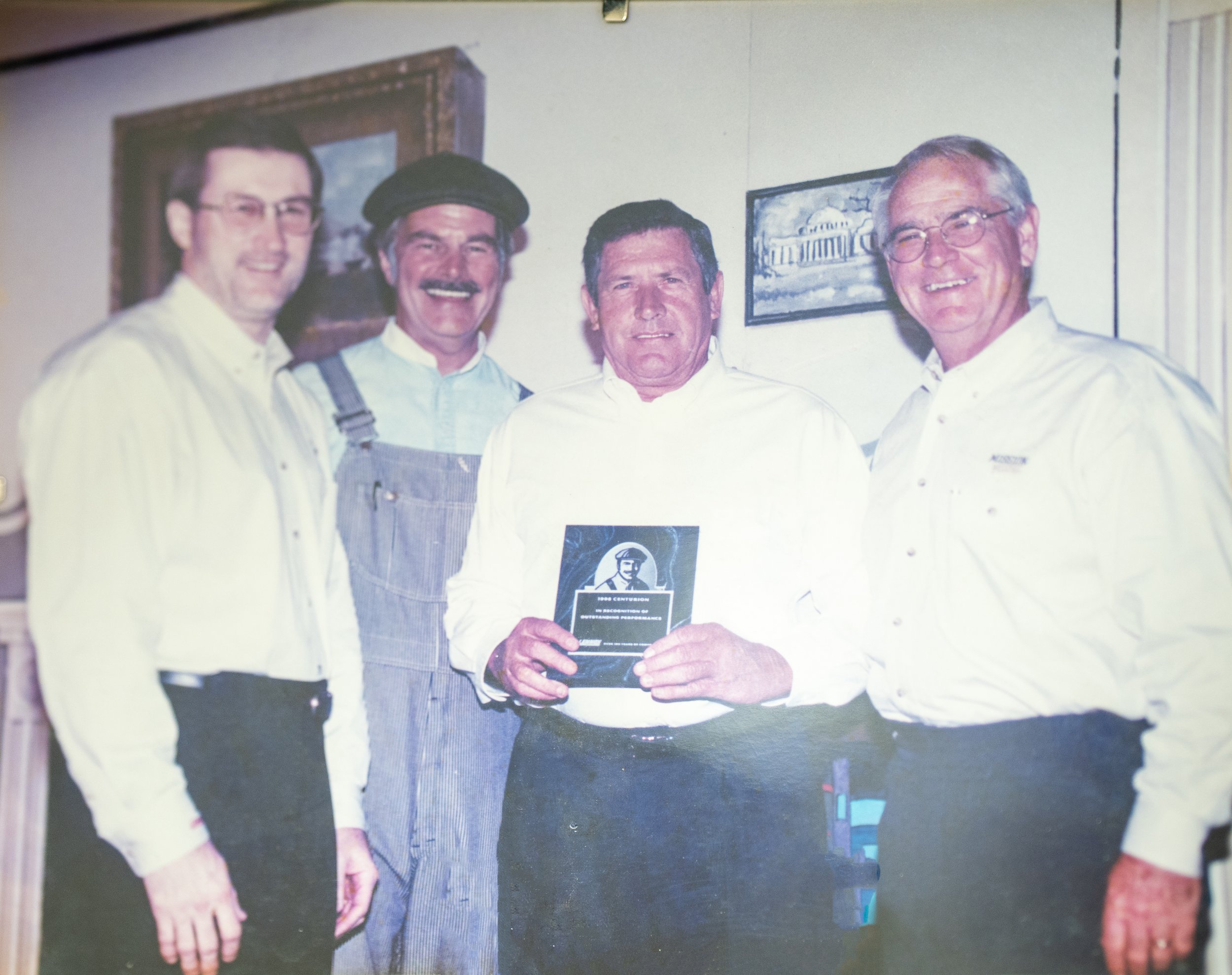 Four men standing together indoors, one holding a plaque or award, with framed pictures on the wall behind them.