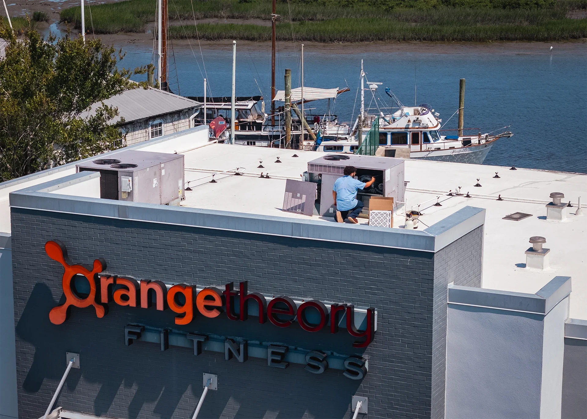 A man working on the rooftop of the OrangeTheory Fitness building with boats docked on water in the background.