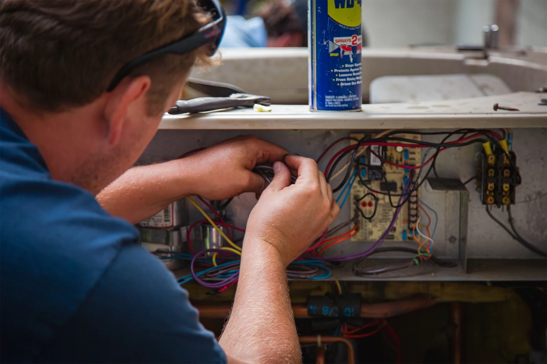 A technician working on electrical wiring inside a panel, with various wires and components visible, and a can of WD-40 on the work surface.