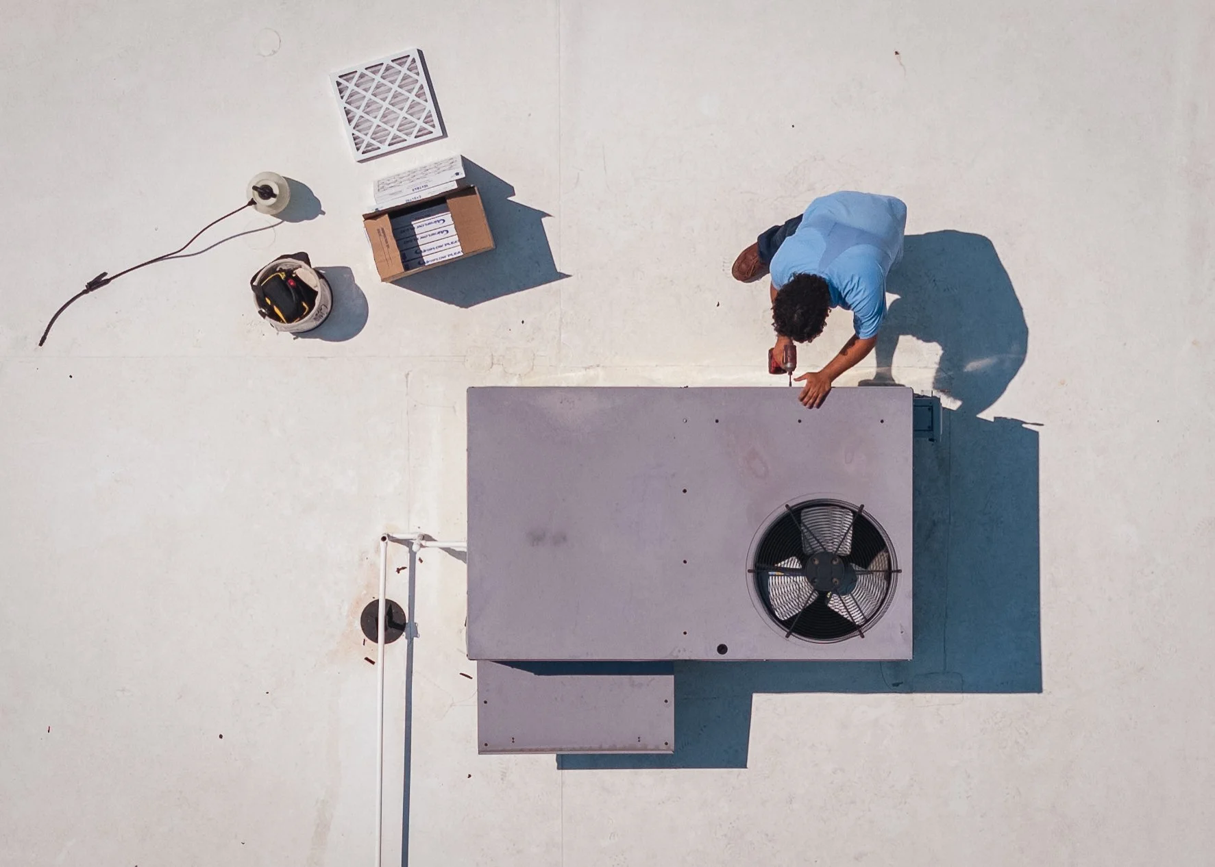 A person working on a rooftop air conditioning unit, using a power drill, with tools and parts nearby.