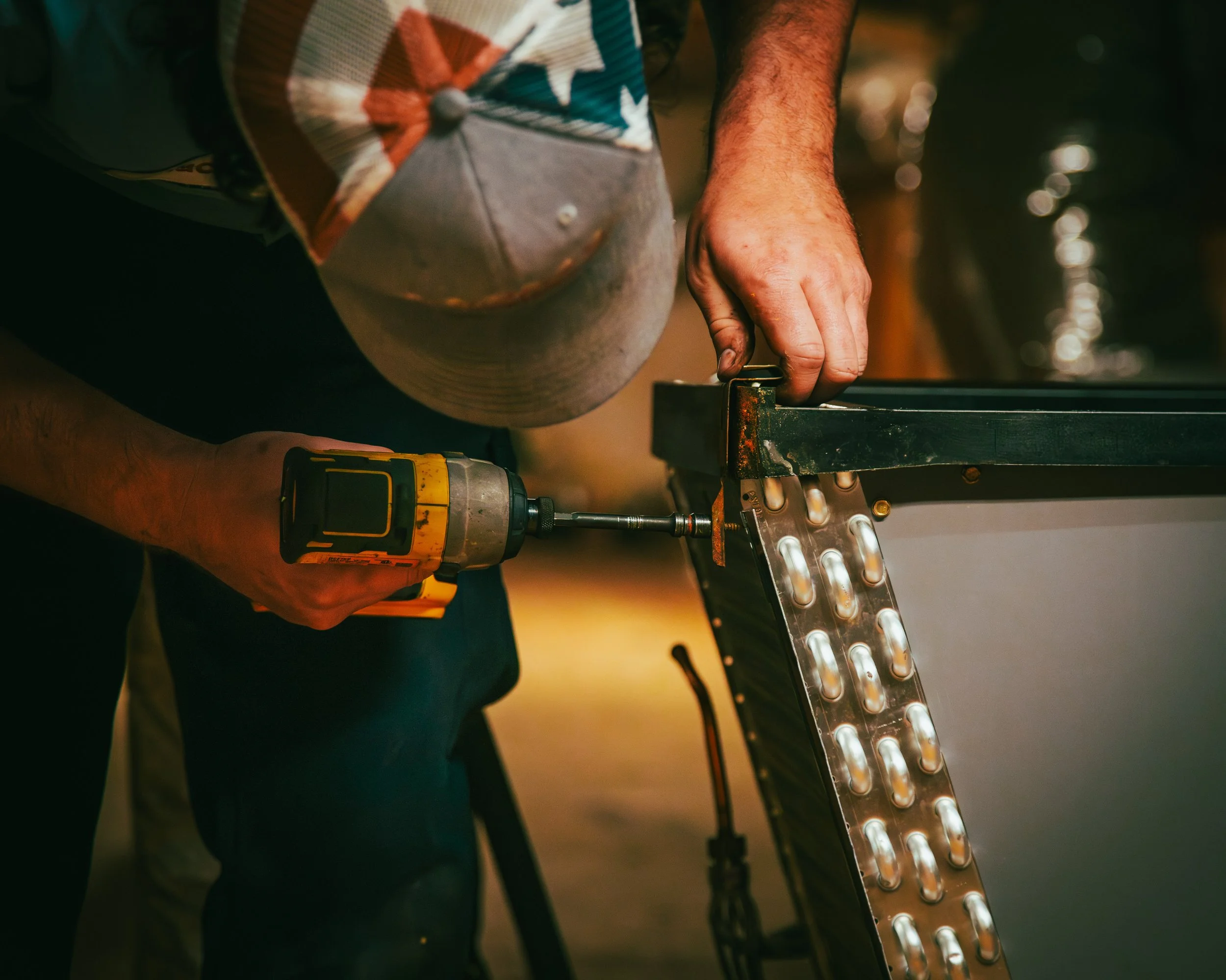 A person uses a drill to work on a metal panel. The person is wearing a colorful cap and has a tattooed arm. The focus is on their hands and the tools they are using.