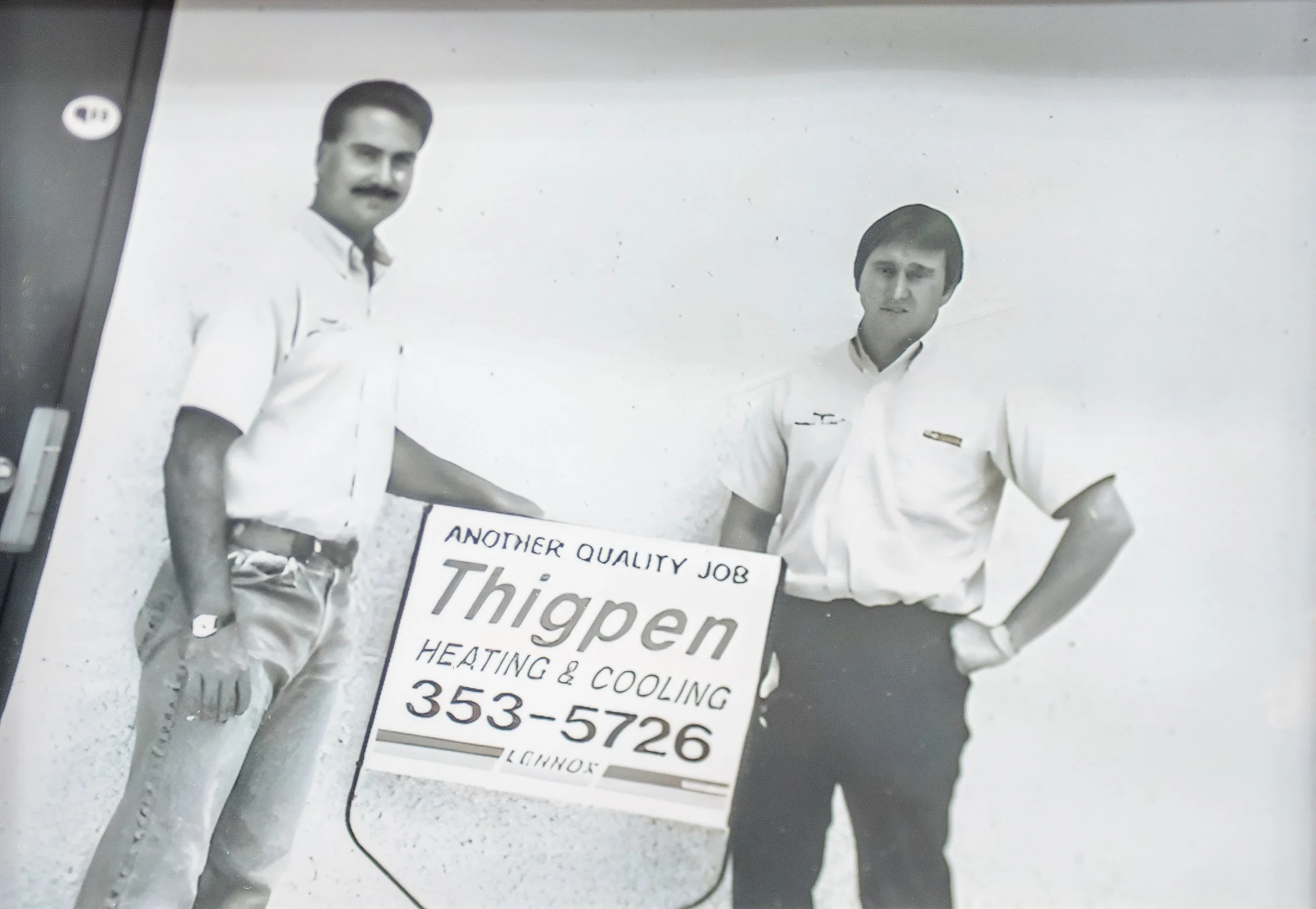 Black and white photo of two men in white shirts standing behind a sign for a heating and cooling company, one with a moustache and the other with a serious expression.