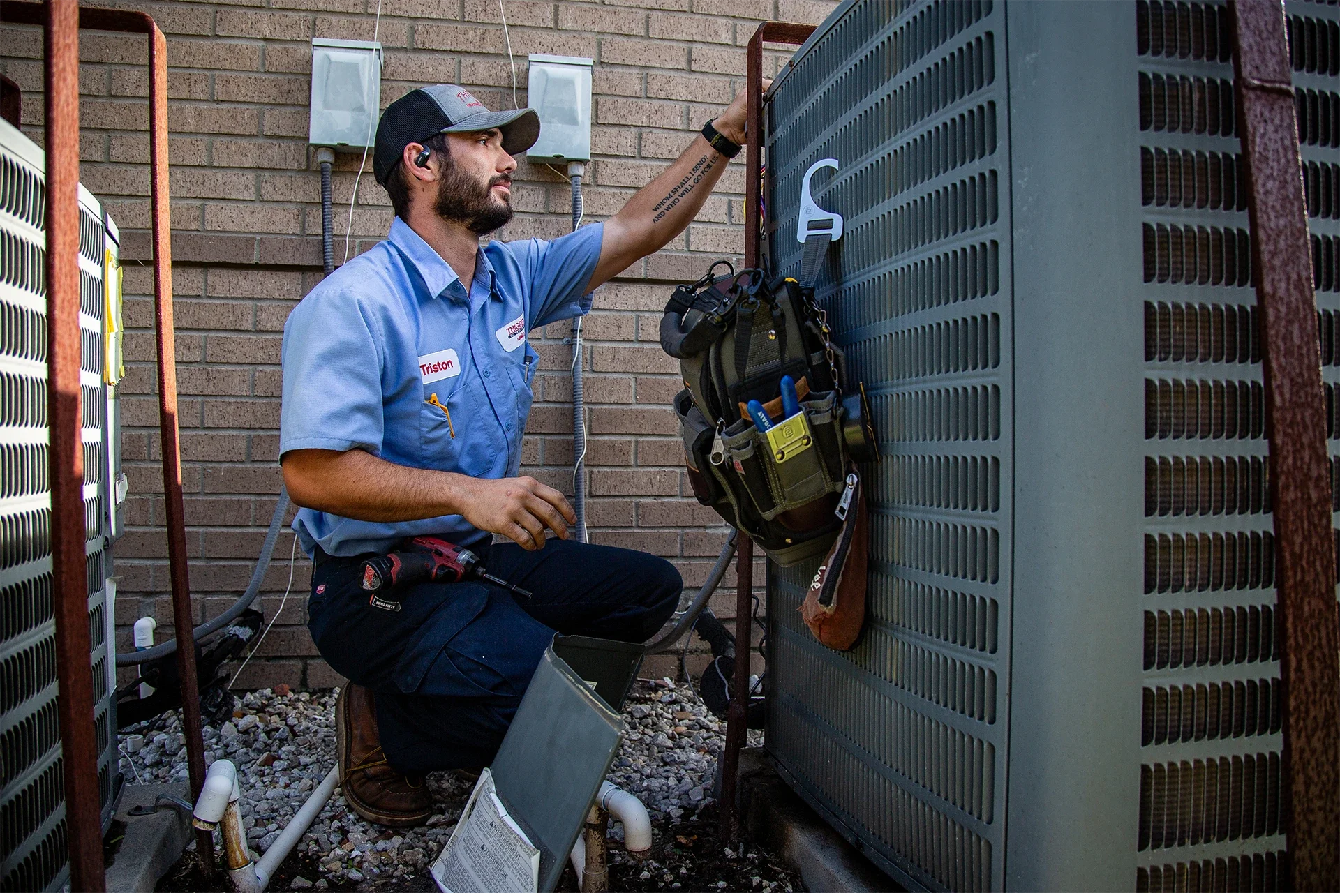A technician kneeling next to an outdoor air conditioning unit, working on it with tools, wearing a blue uniform shirt, a cap, and a tool belt, set against a brick wall.