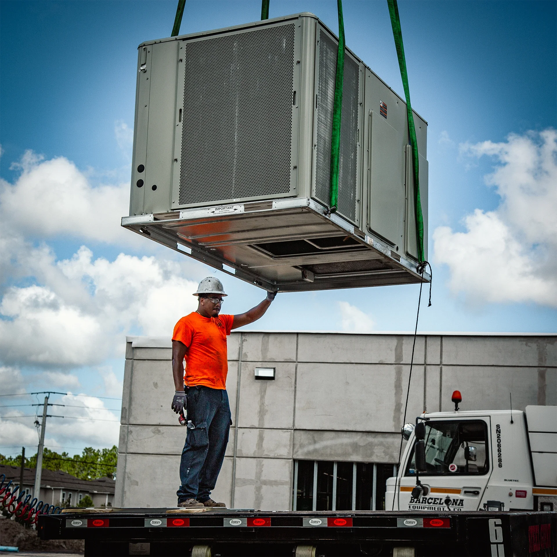 Worker in an orange shirt and white hard hat guiding a large HVAC unit being hoisted by a crane on a flatbed truck.