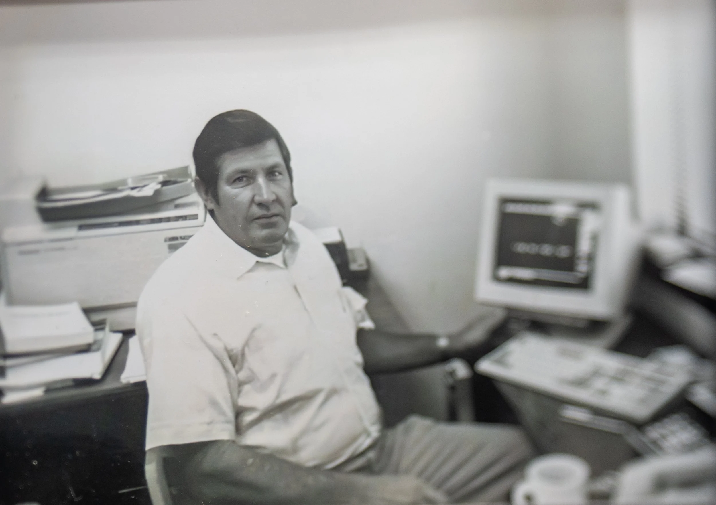 A man with dark hair seated at a desk amidst stacks of paper and electronic equipment in a workspace.