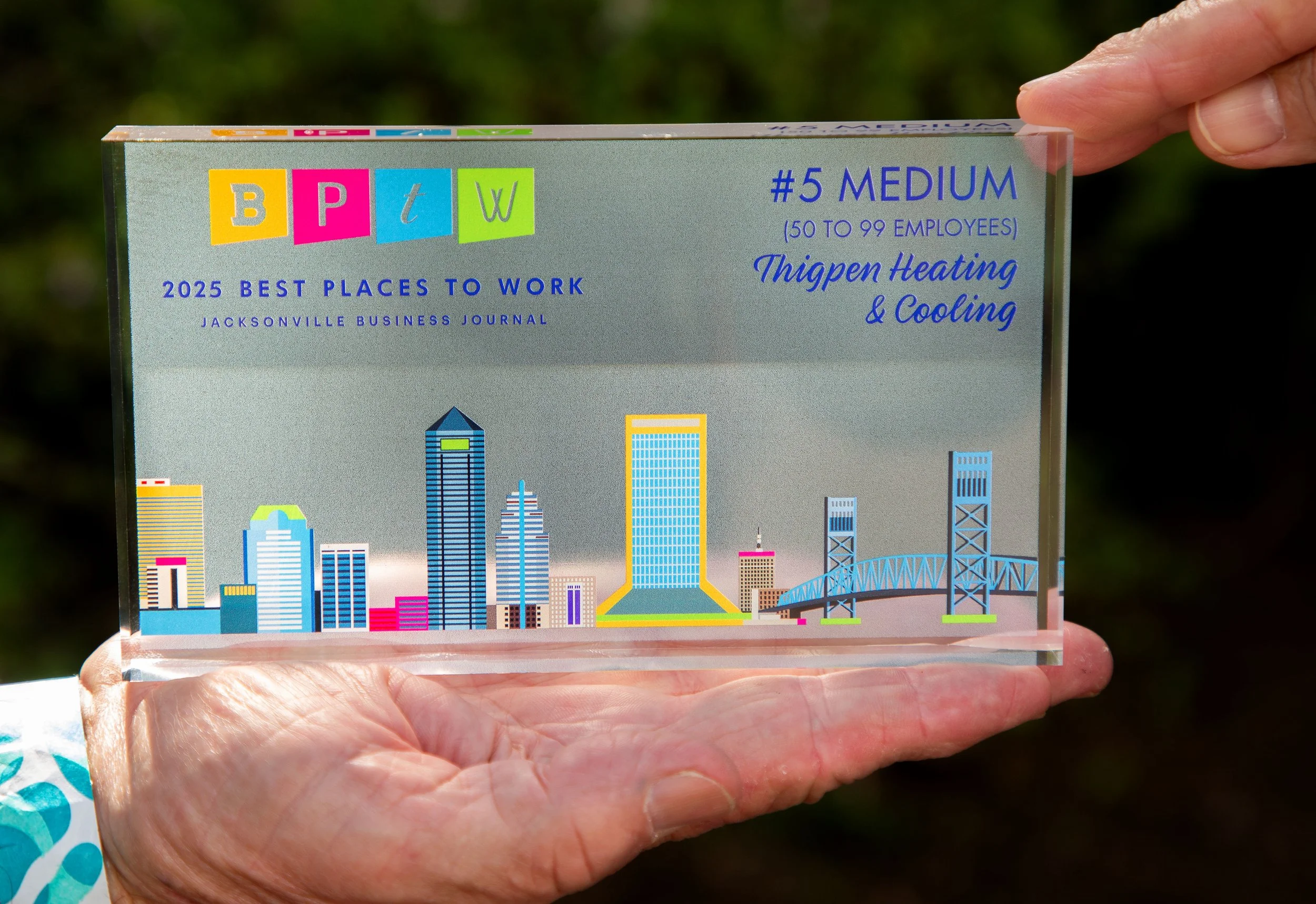 Close-up of a clear acrylic award plaque with a colorful cityscape design at the bottom, being held in someone's hand outdoors. The plaque features text about Jacksonville's best workplaces in 2025, with rankings and category details.