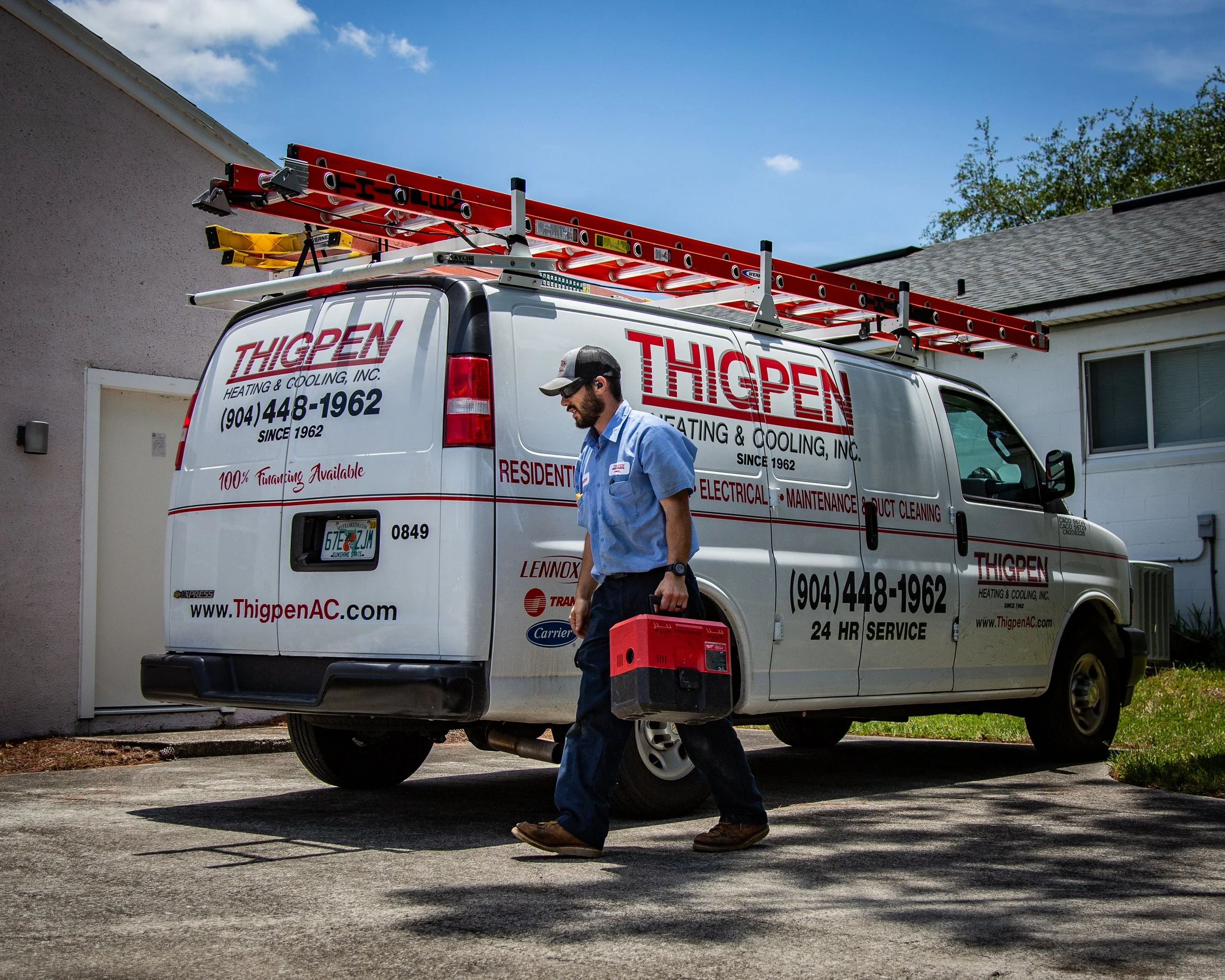 A technician walks past a service van with the company name 'Thigpen Heating & Cooling, Inc.' parked outside a house, carrying a red toolbox, with a ladder mounted on the roof of the van.