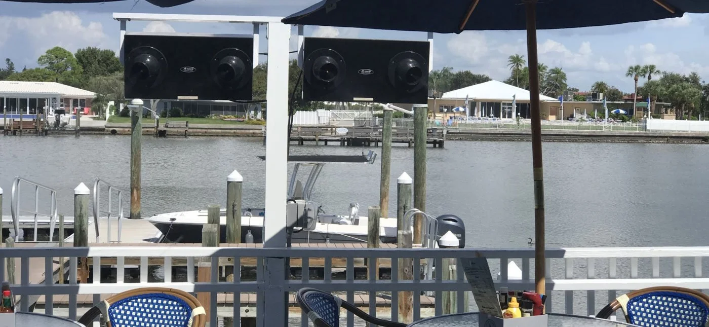 Outdoor waterfront restaurant seating area with a view of a dock, boat, and water, featuring a large black speaker system, an umbrella, and tables with seating.