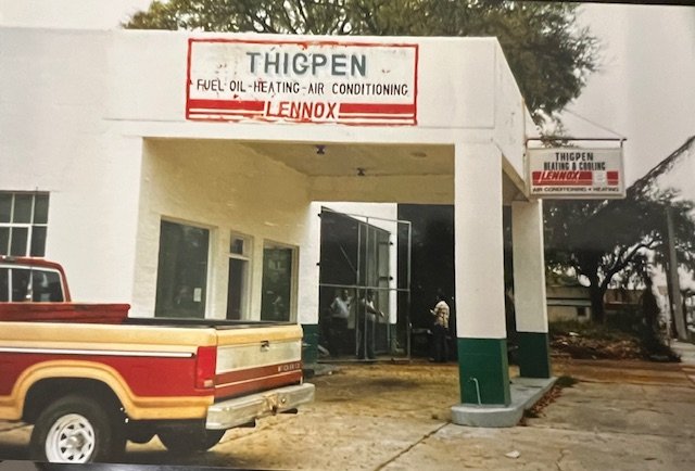 Exterior of Thigpen heating and cooling business with a white building, a sign listing services, a red and beige truck parked nearby, and a tree in the background.