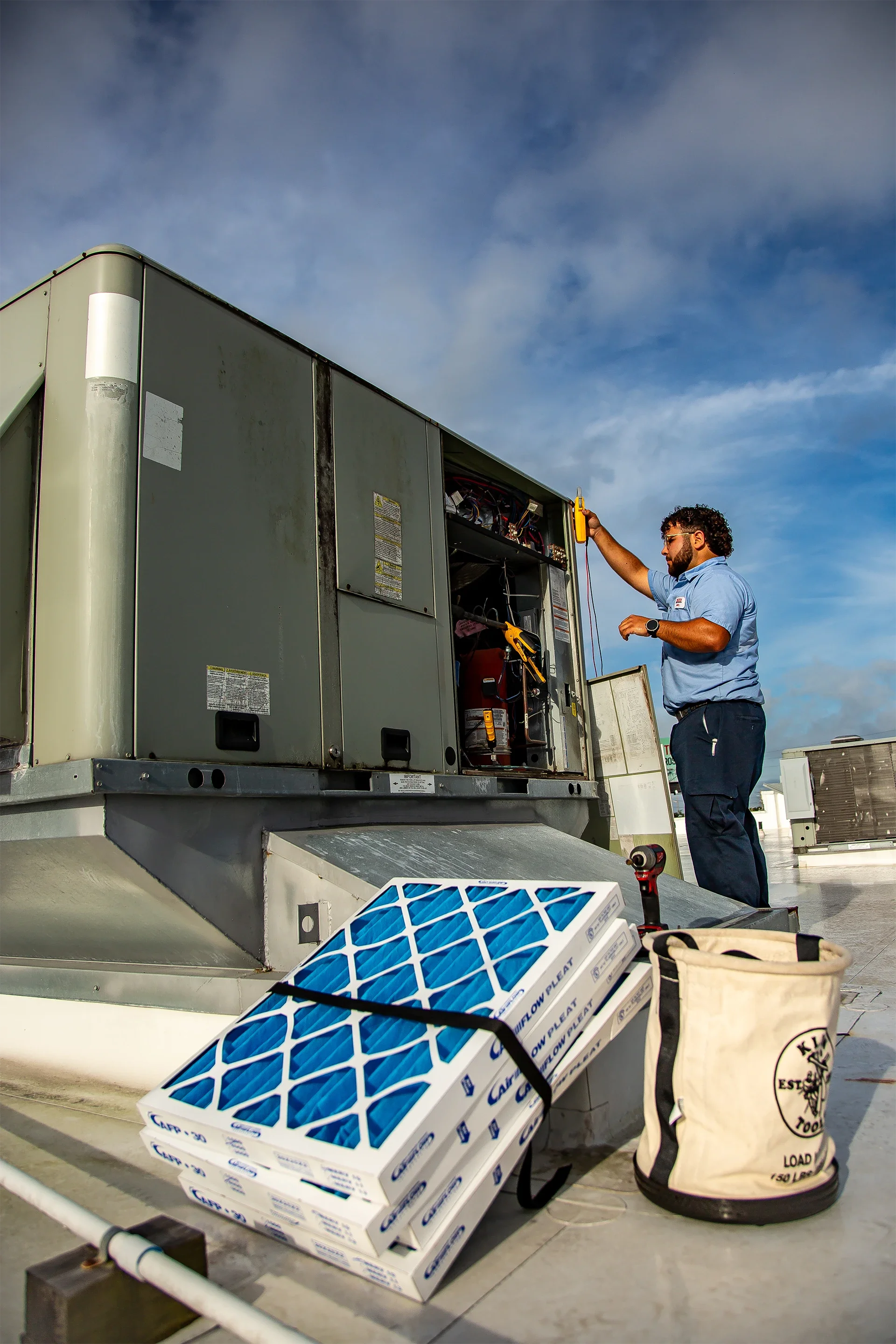 A technician working on an HVAC unit on a rooftop, using tools and wearing safety gear, with parts and equipment nearby.