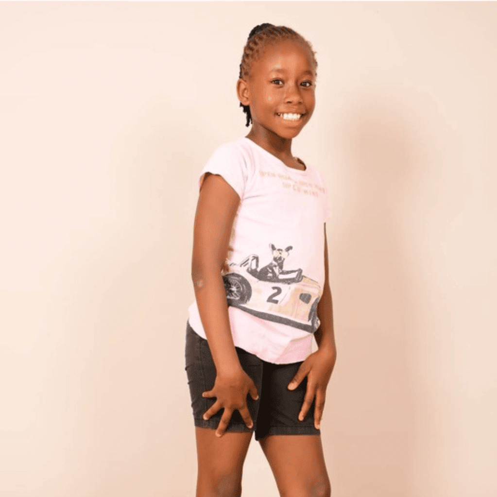 A young girl with braided hair smiling, wearing a white t-shirt with a racing car graphic and black shorts, standing against a plain light-colored wall.
