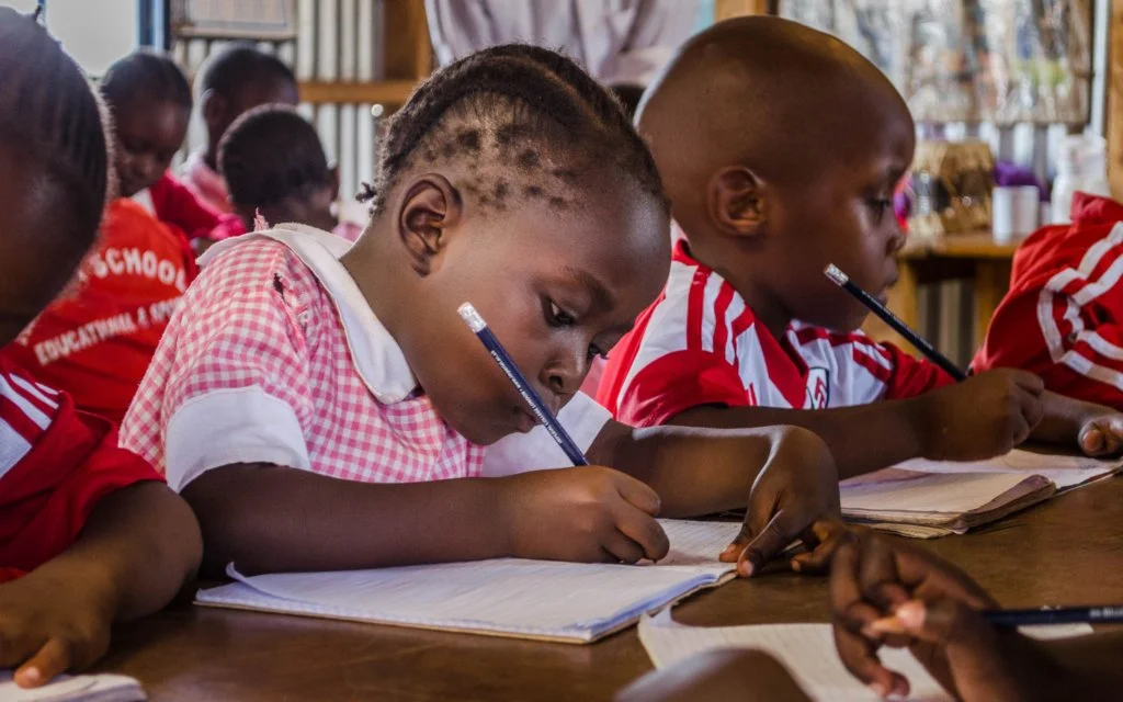 Young children sitting at desks in a classroom, focused on writing in notebooks with pencils, wearing school uniforms.