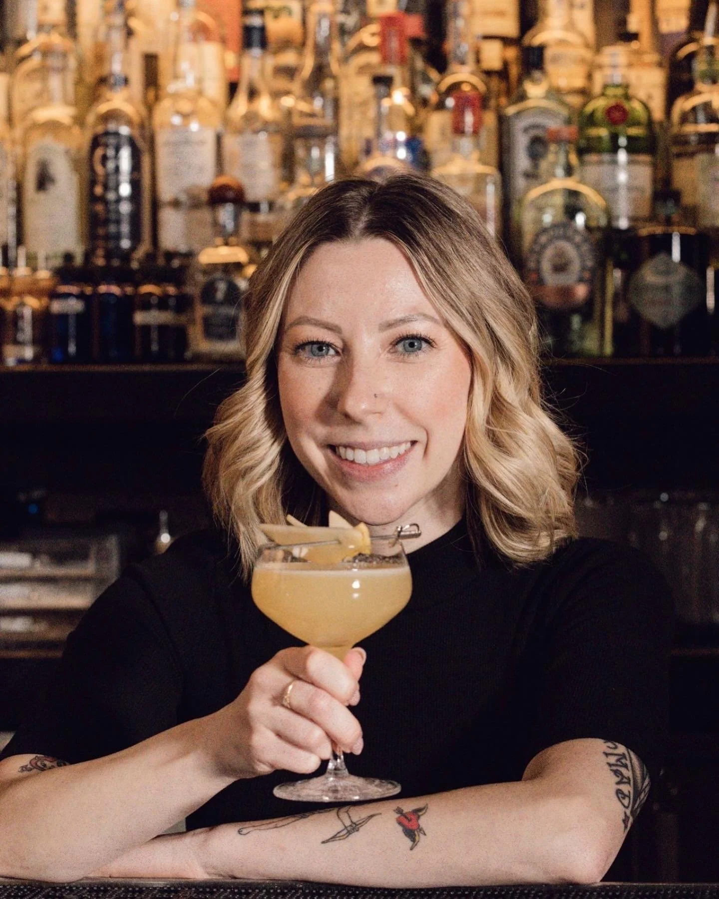 A smiling woman with blonde hair and tattoos on her arms is holding a cocktail glass with a yellow drink and a garnish, sitting at a bar with bottles and wooden shelves in the background.
