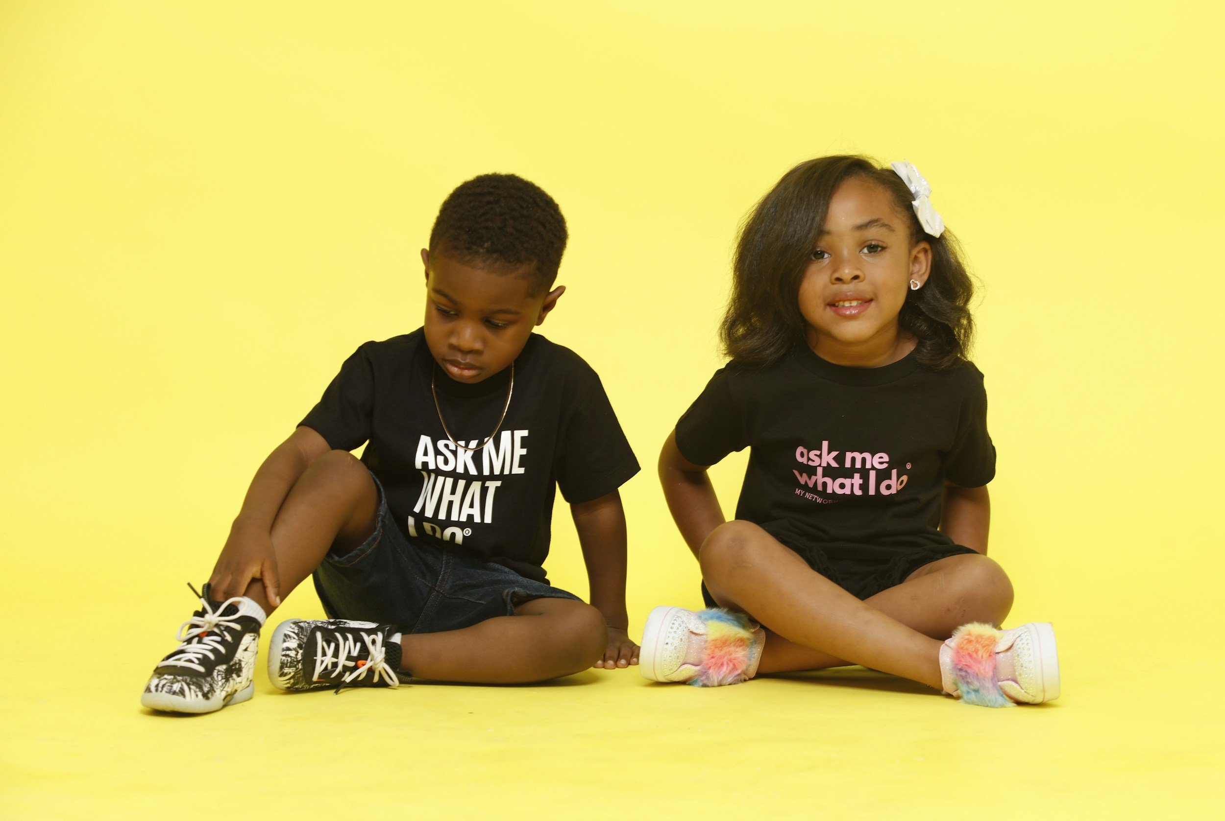 Two children sitting on the floor against a yellow background, wearing black T-shirts with pink and white text.