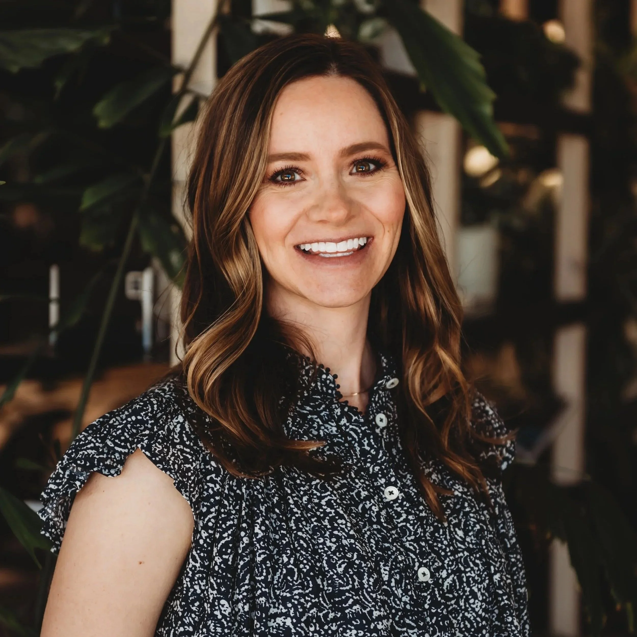 A woman with brown hair styled in loose waves, wearing a patterned black and white blouse, smiling in front of a leafy background.