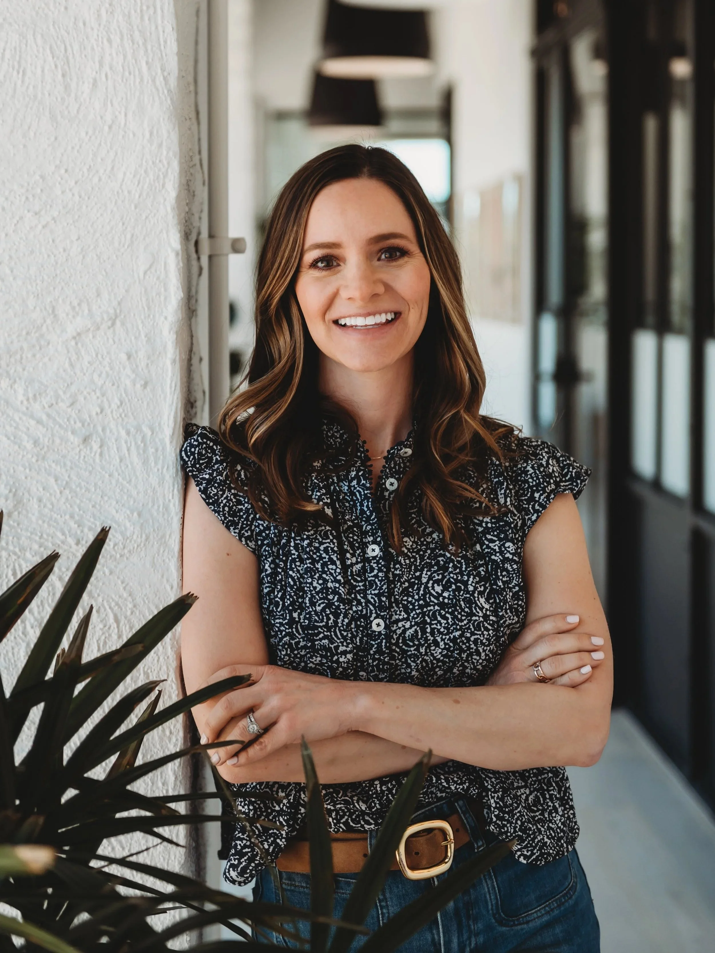 Smiling woman with shoulder-length brown hair, wearing a black patterned sleeveless blouse, standing with arms crossed indoors next to a white textured wall and a plant.