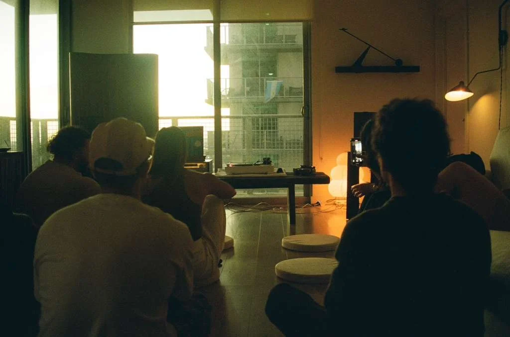 People sitting indoors watching a screen during a video or presentation, with bright light from a window behind them.
