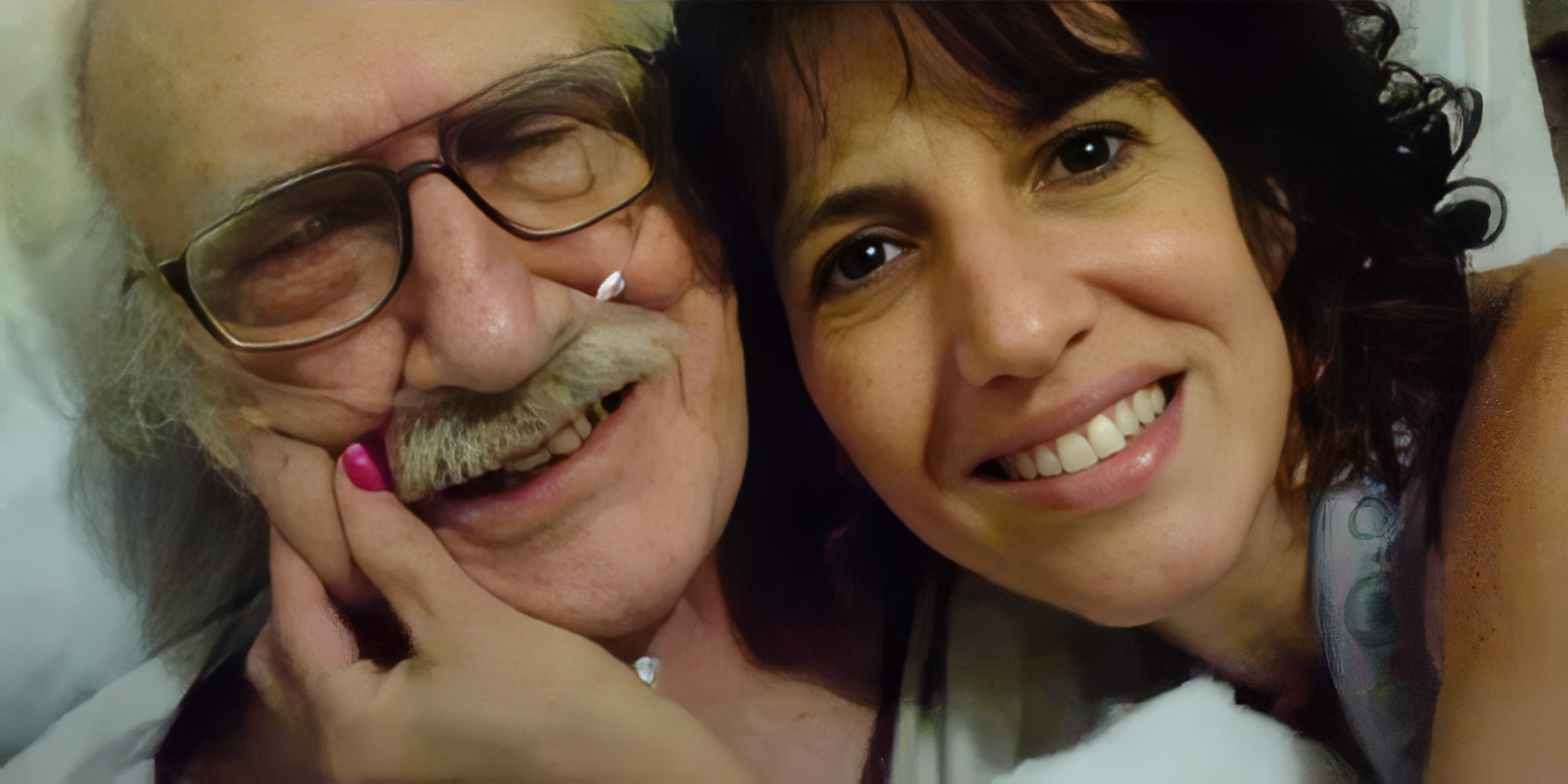 Close-up portrait of a smiling elderly man with glasses and a mustache and a young woman with curly dark hair, both lying down close together, smiling at the camera.