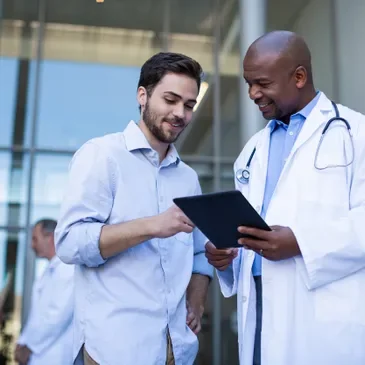 Two men, one in a white lab coat with a stethoscope, and one in casual clothes, looking at a tablet together outside a medical building.