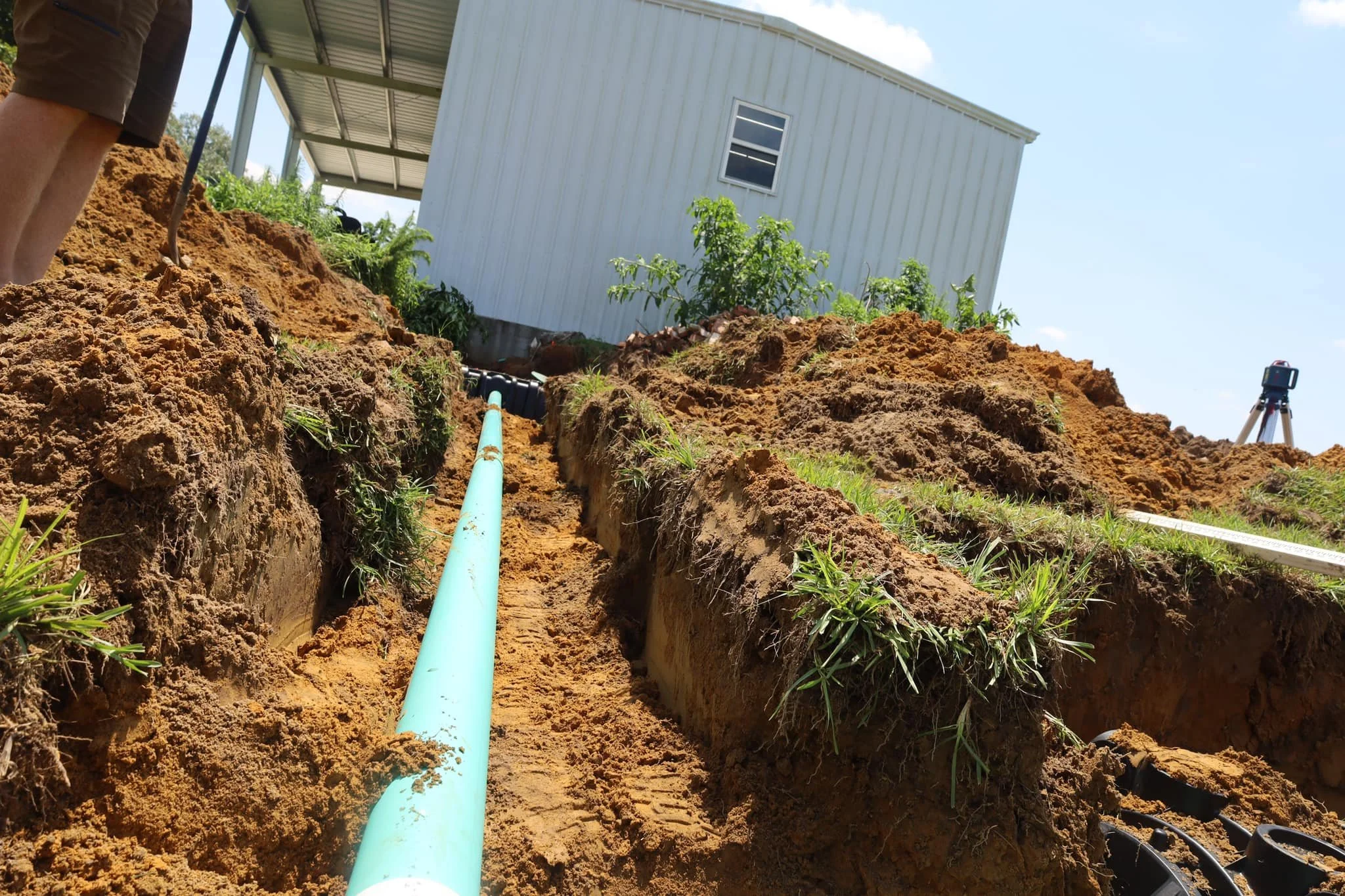 A construction site showing a trench with a blue pipe installed, neighboring a white metal building, and an instrument on a tripod in the background, under a partly cloudy sky.