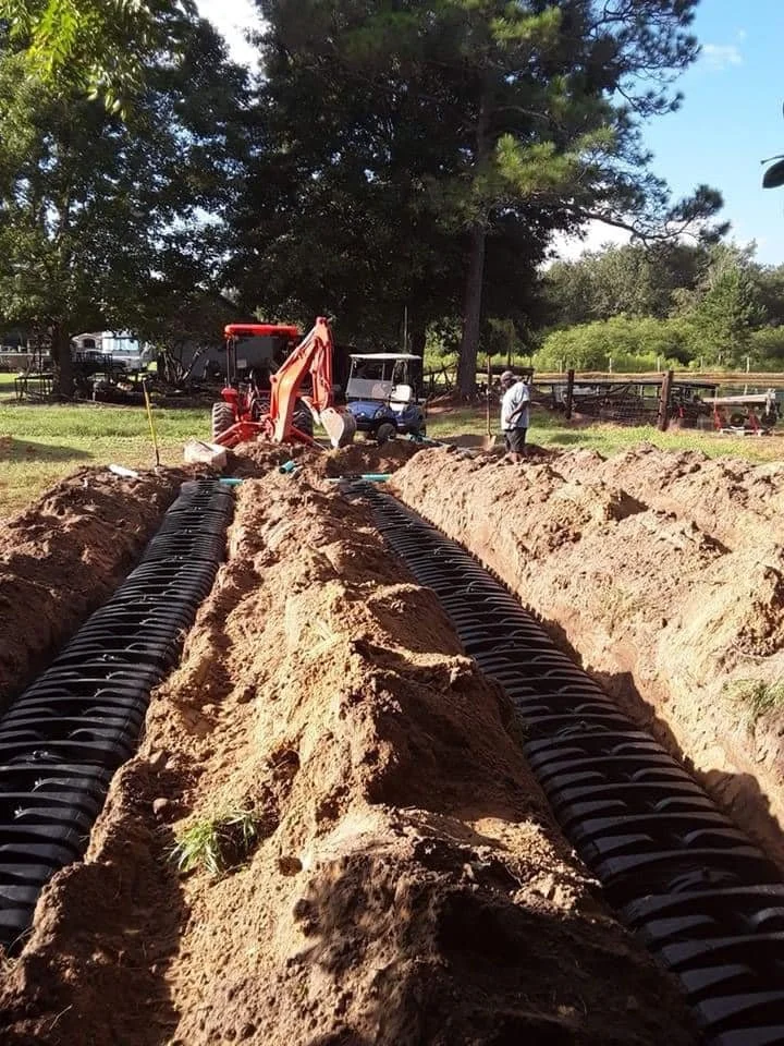Excavation site with trenches being dug for underground piping, surrounded by trees and construction equipment, including a small orange backhoe and a golf cart.