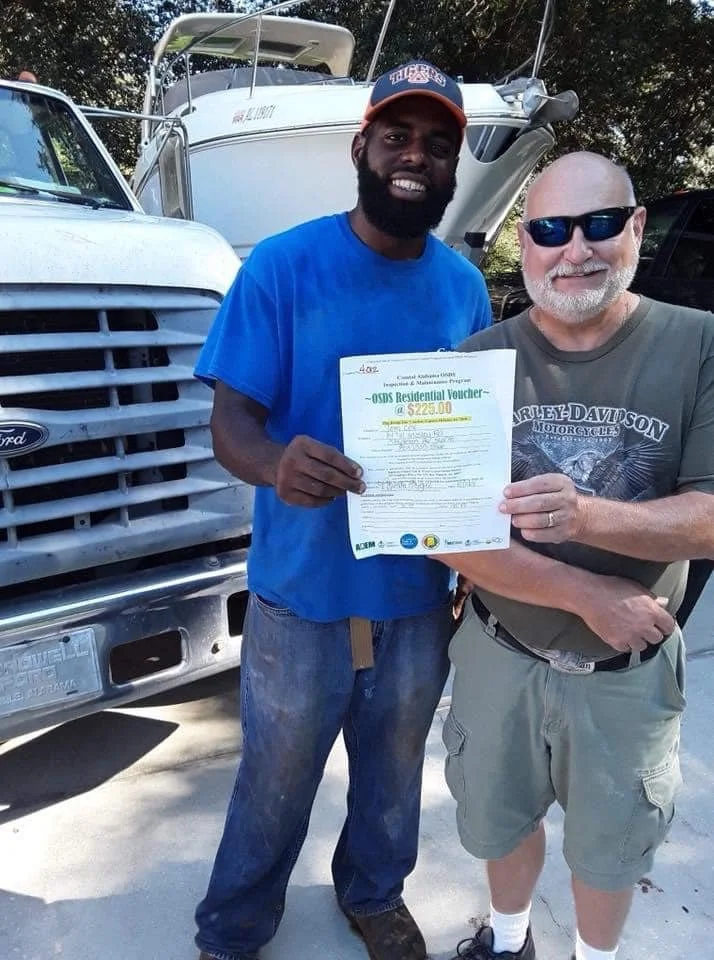 Two men standing outdoors, smiling and holding a document. One man has a dark complexion, beard, and is wearing a blue t-shirt and an orange cap. The other man has light skin, a beard, sunglasses, and is wearing a gray t-shirt with a Harley Davidson 