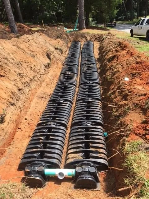 Two black corrugated pipes laid parallel in a trench at a construction site surrounded by dirt, with trees and parked cars in the background.