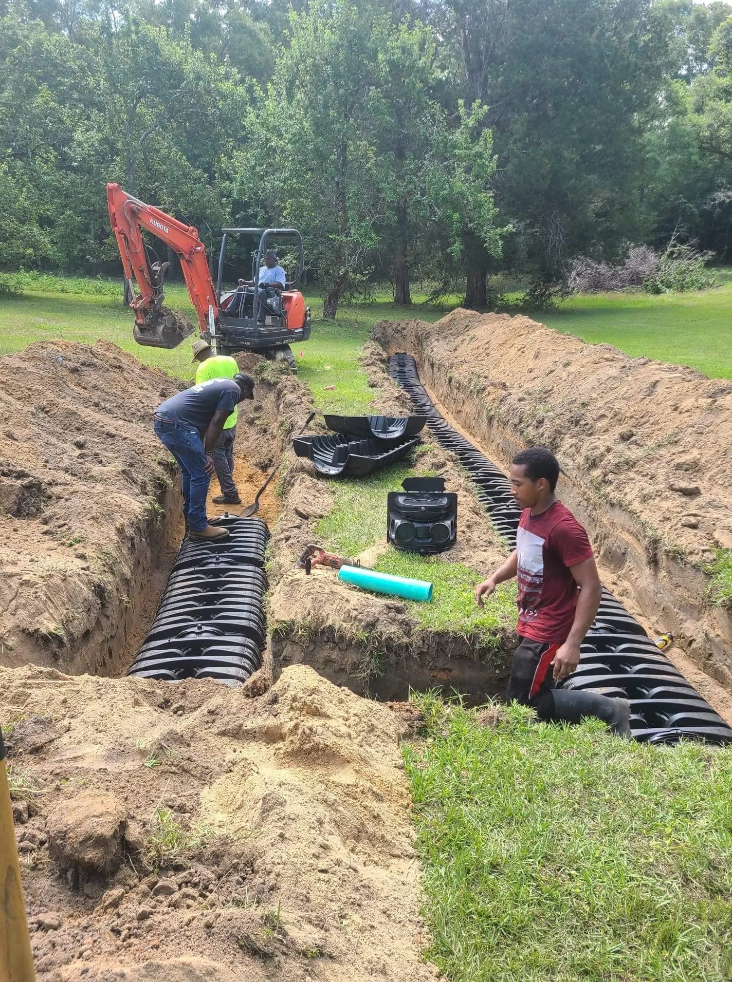 Workers installing large black drainage pipes in a partially dug trench in a grassy area, with a small excavator and trees in the background.
