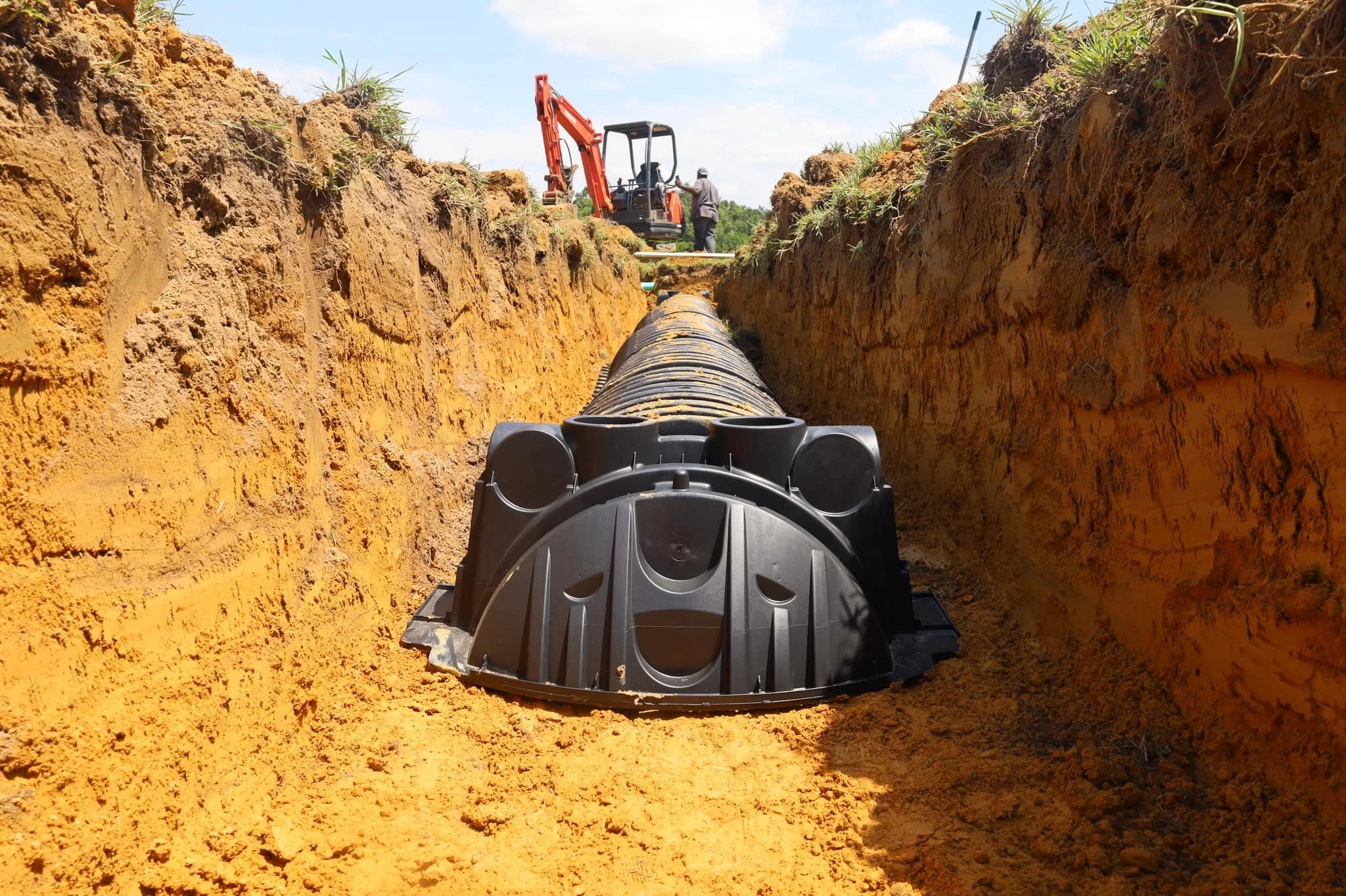 View from the back of a vehicle traveling through a dirt trench with orange soil, with a small excavator and a person in a hat working at the top edge of the trench, and a pipe running along the bottom of the trench under the vehicle.
