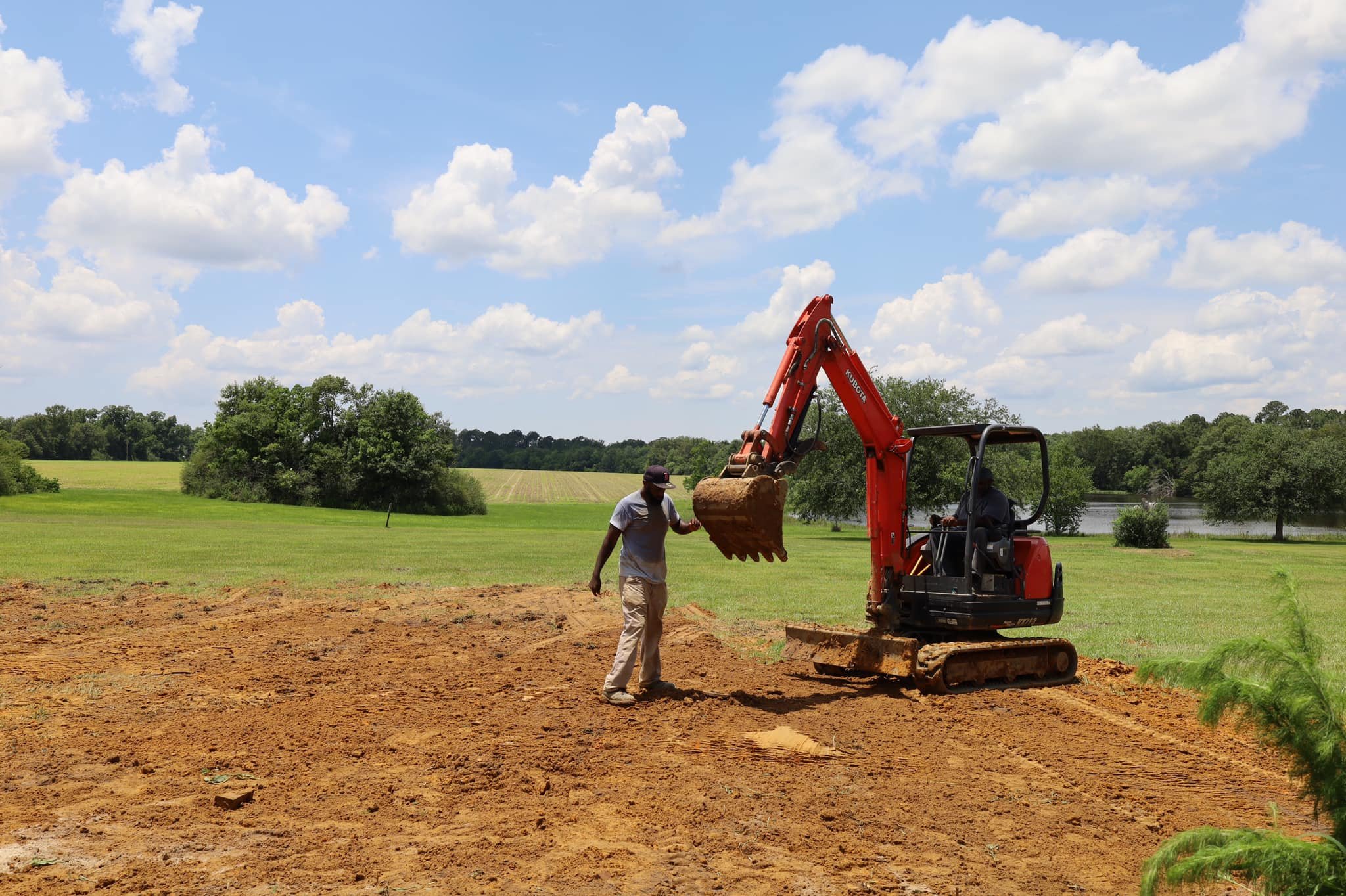 Two men working outdoors with a small orange excavator on a dirt patch, green fields and trees in the background under a partly cloudy sky.