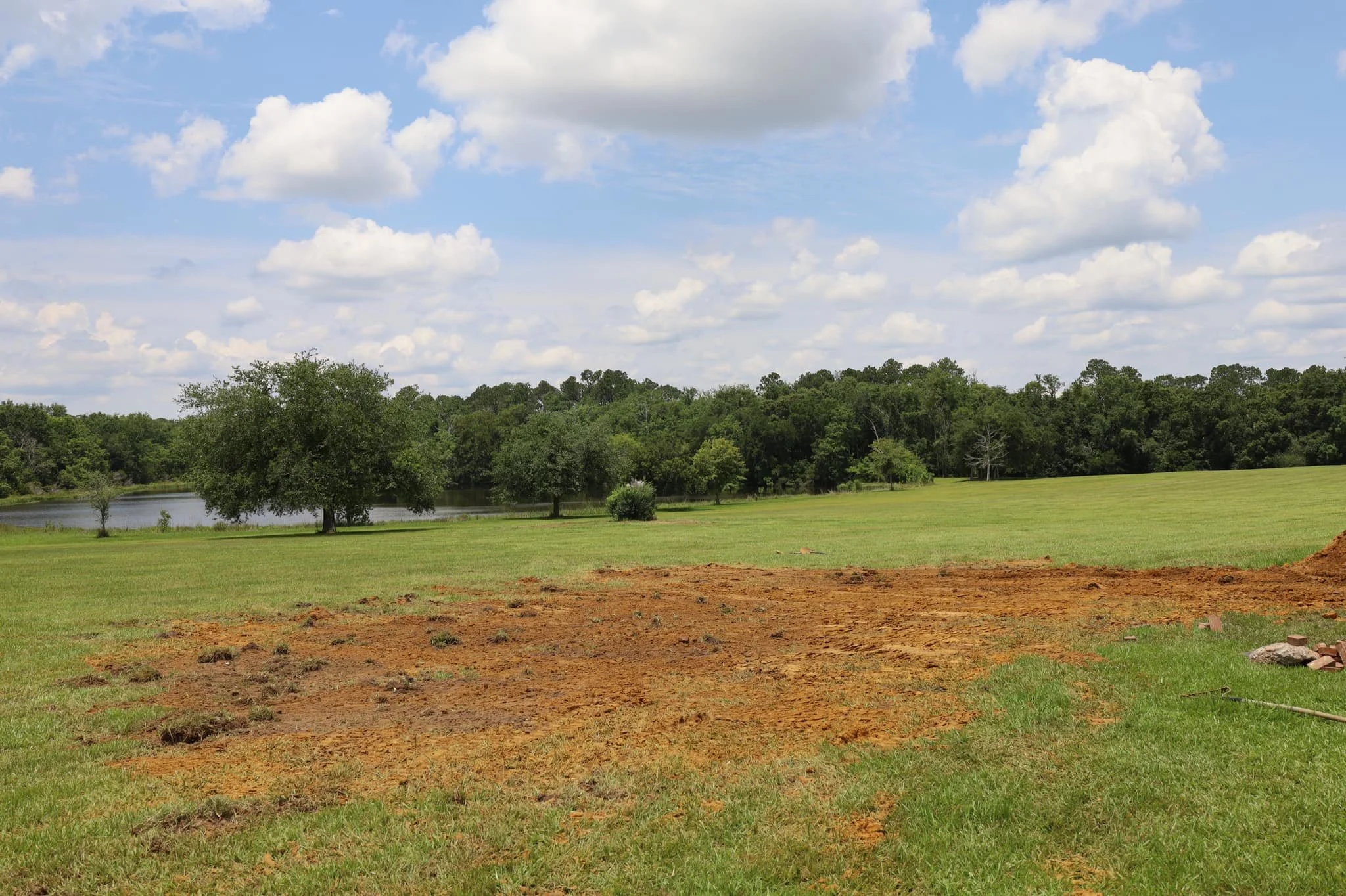 A large grassy field with patches of dirt and some trees near a body of water, under a partly cloudy sky.