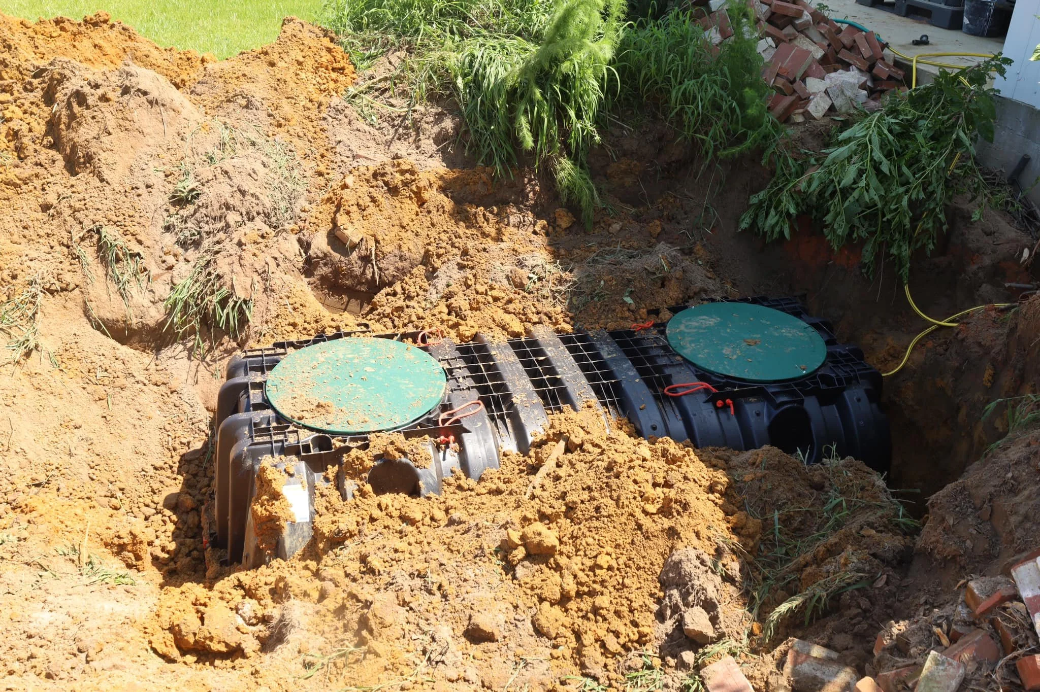 A trench dug in the ground with dirt piled on the side, surrounded by grass and plants. There are two black underground utility telecommunication and electrical boxes with green covers in the trench, connected to yellow cables. Construction bricks and tools are visible in the background.