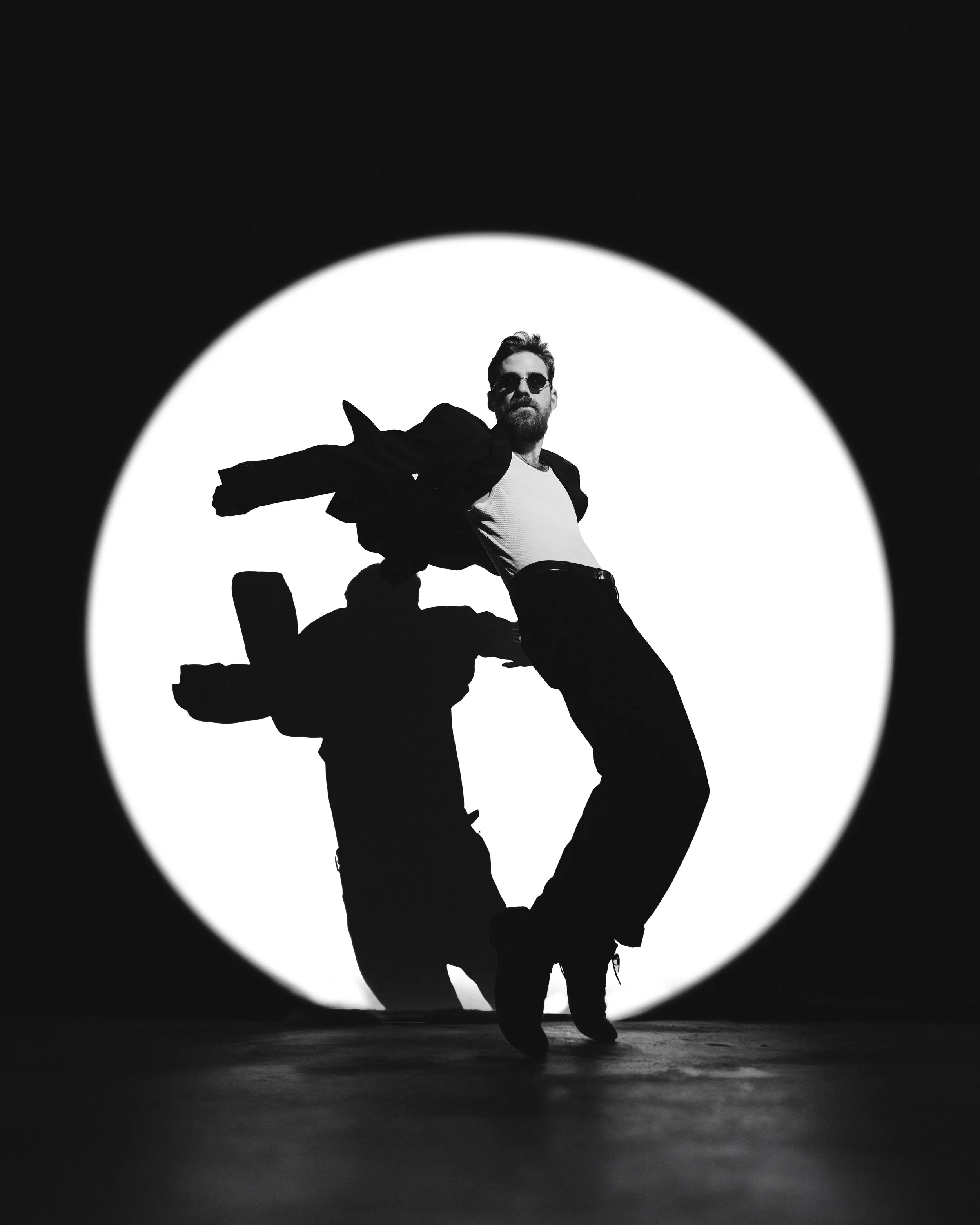 A man with a beard and sunglasses poses with one leg bent and the other straight, wearing a black jacket, light shirt, dark pants, and shoes, against a large circular white background in black and white.