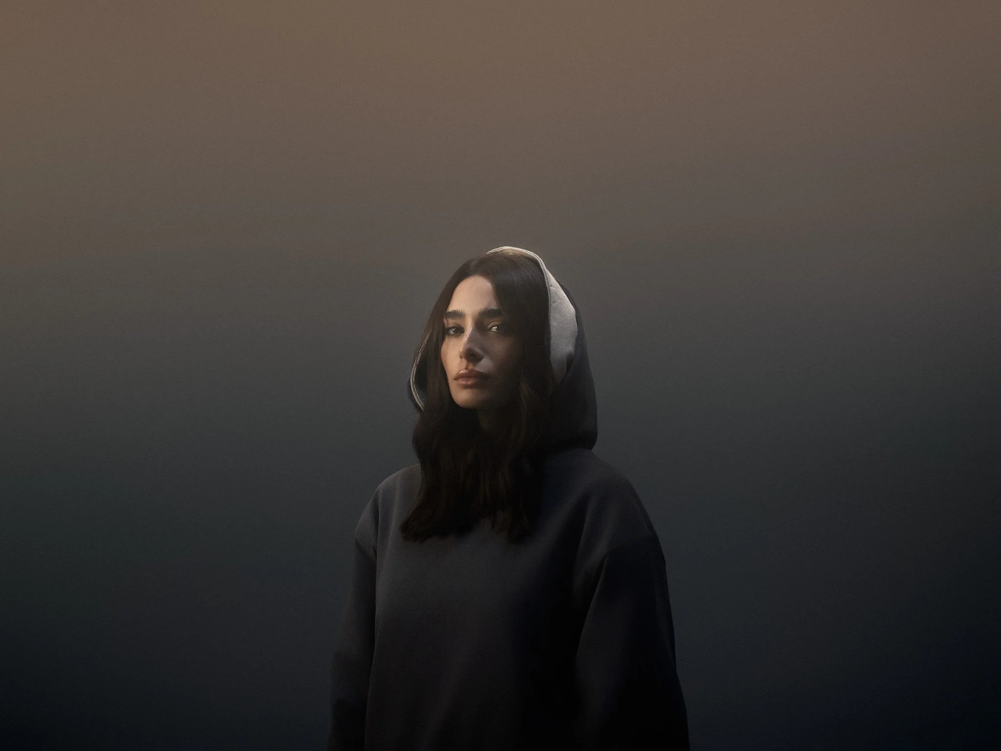 A woman with dark hair and a hoodie standing against a dark gradient background