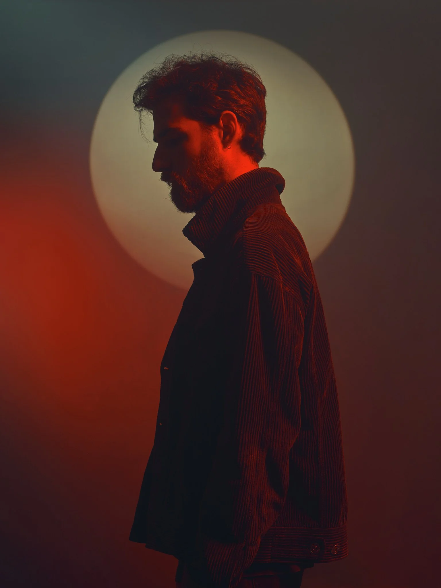 Silhouette of a man with a beard and hair styled back, looking down, standing in front of a bright circular light, with red and orange lighting effects.