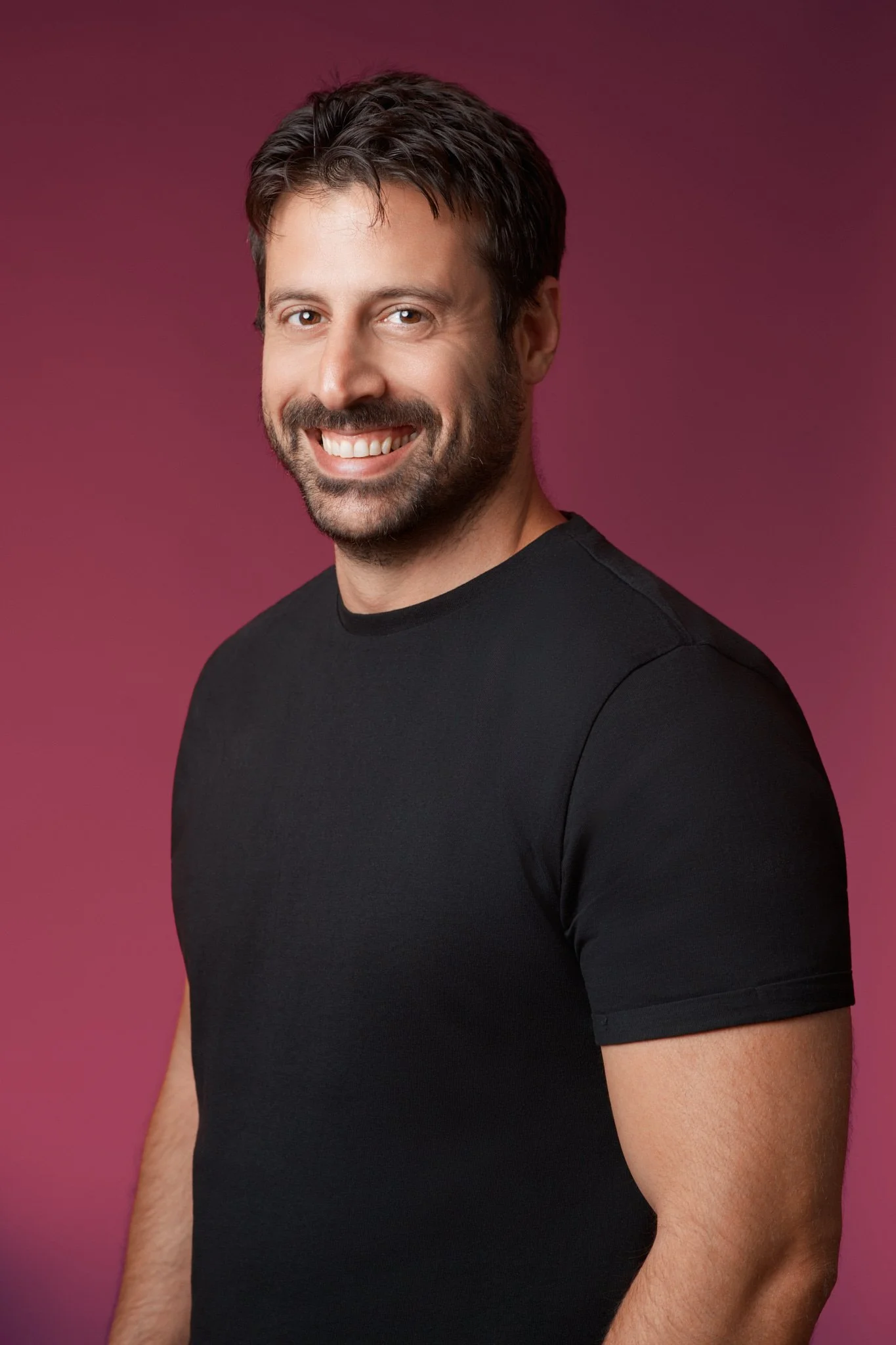 A smiling man with dark hair, beard, and mustache wearing a black t-shirt against a pinkish-purple background.