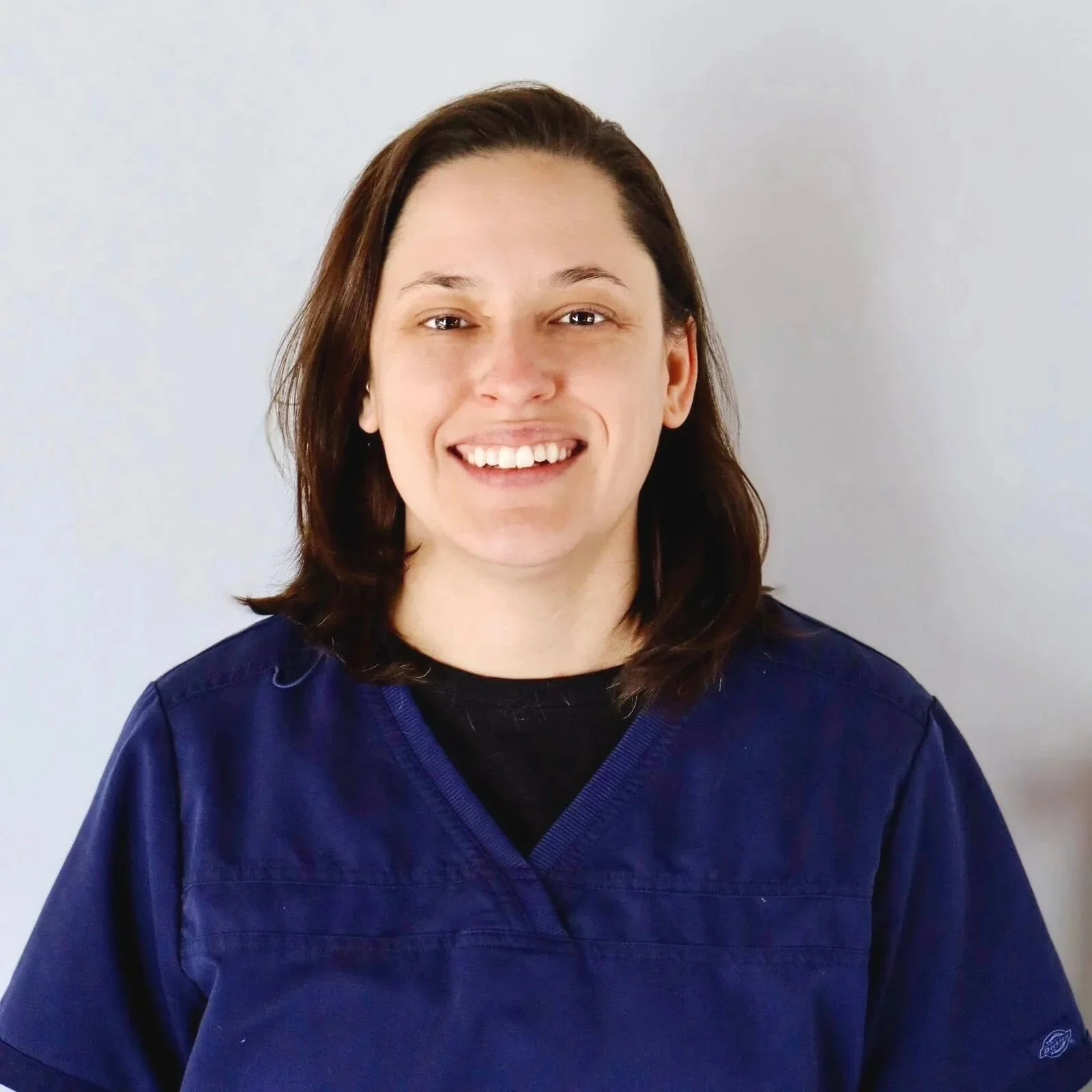 A woman with brown hair, wearing navy scrubs, smiling and standing against a plain light gray background.