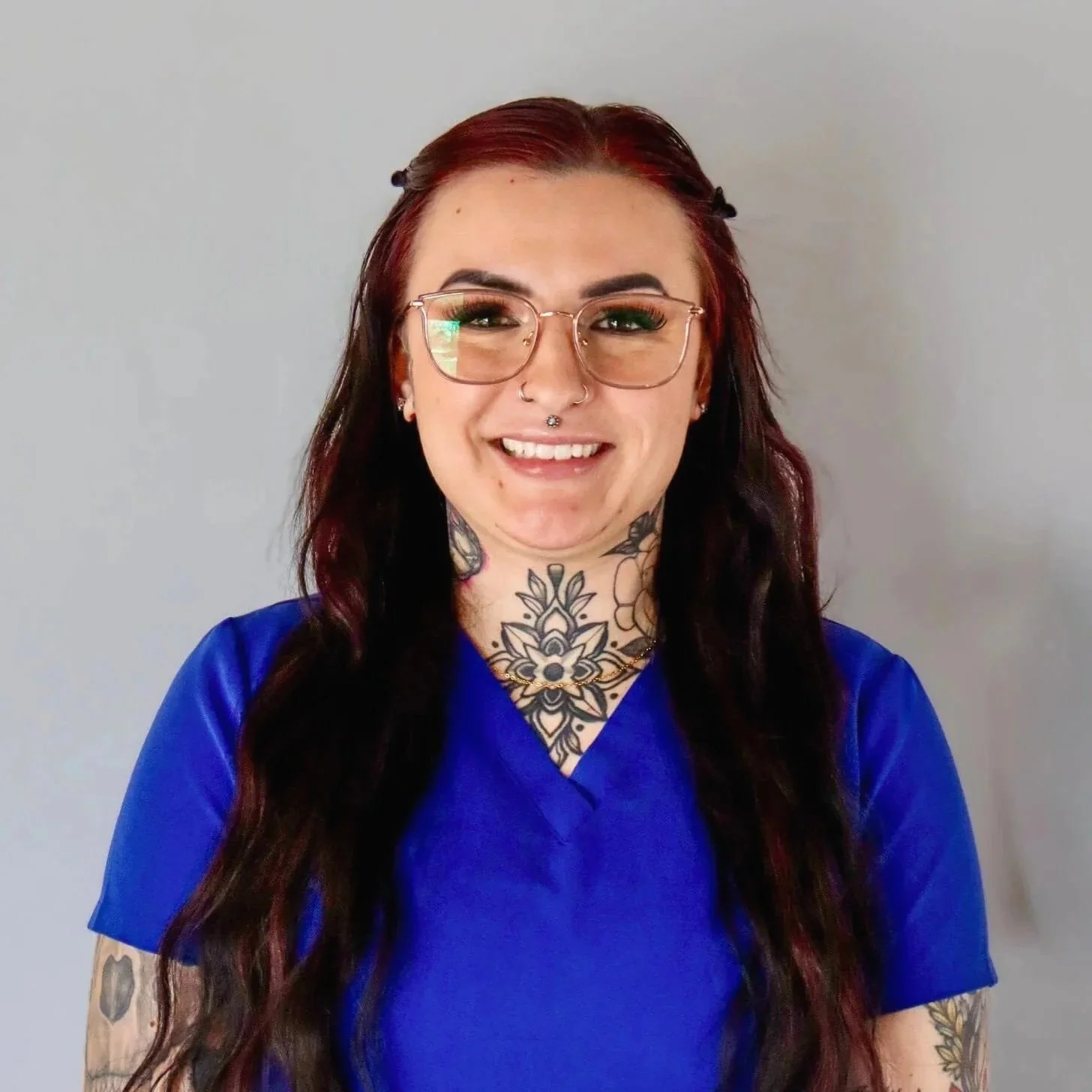 Smiling woman with long wavy red and black hair, tattoos on her neck and arms, wearing glasses and a blue medical scrub top, standing against a light gray background.