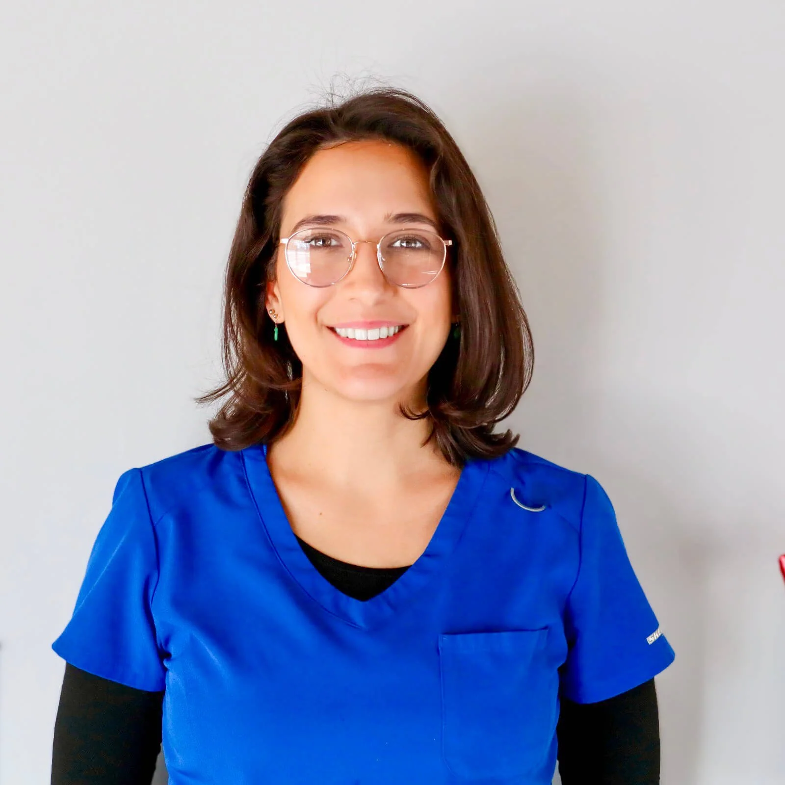 A smiling woman with glasses wearing blue scrubs standing against a plain light-colored wall.