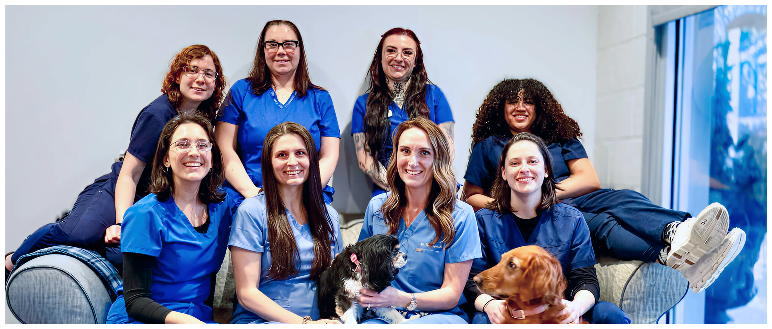 Group of nine women wearing scrubs, posing happily with two dogs in a room with a white wall and a glass door.