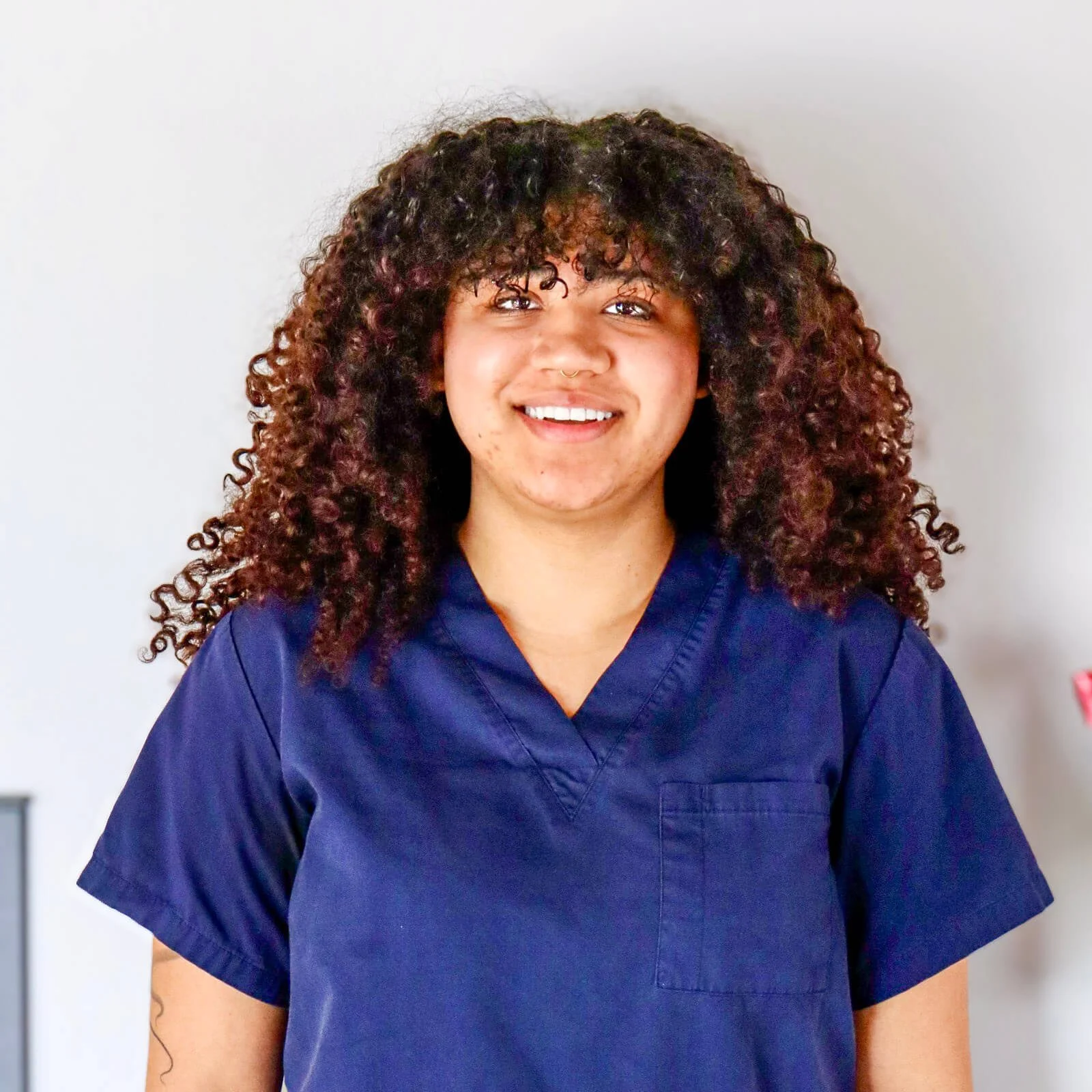A smiling woman with curly hair wearing a navy medical scrub top standing against a plain light-colored wall.