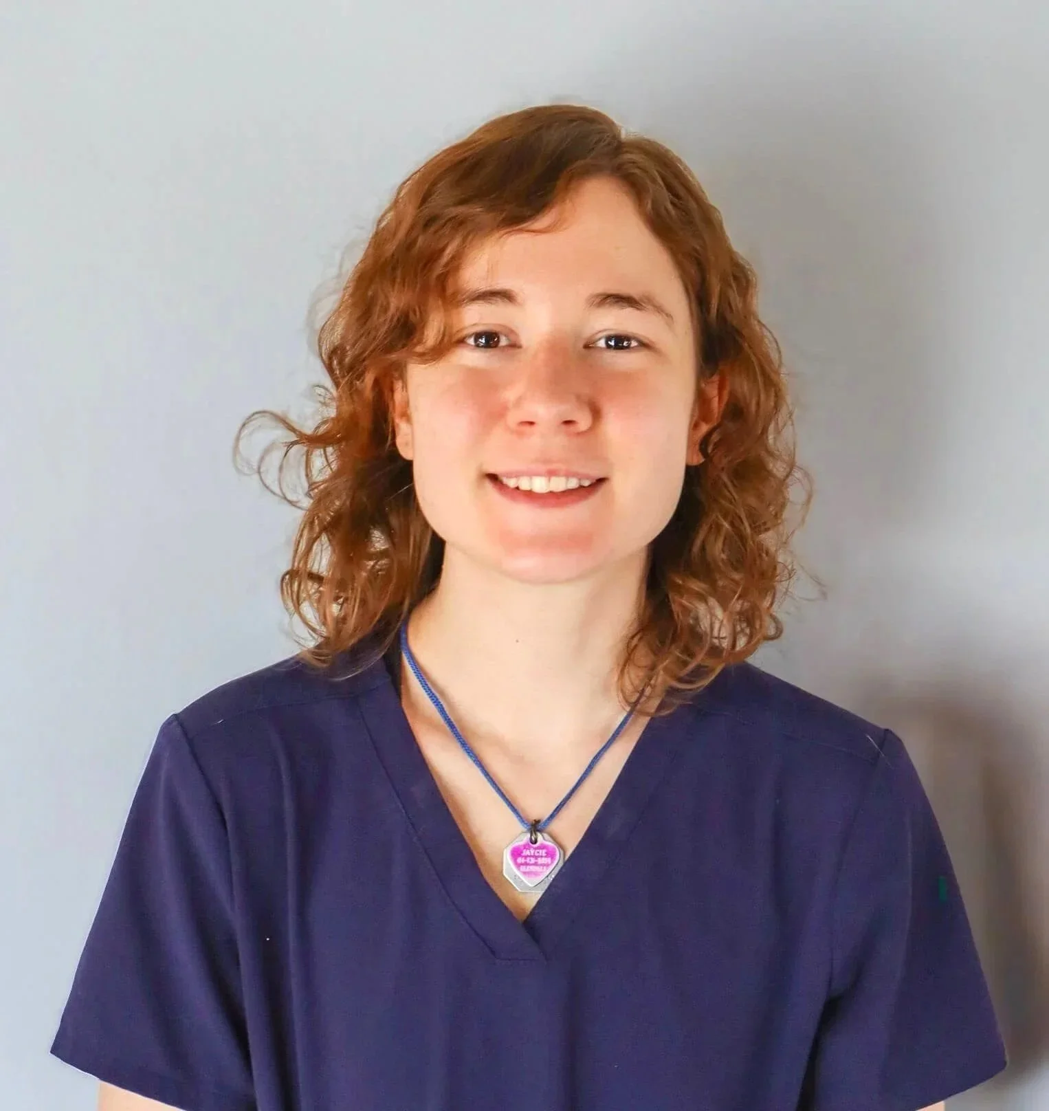 A woman with curly red hair and fair skin, wearing navy scrubs and a purple necklace with a badge pendant, smiling against a plain light gray background.