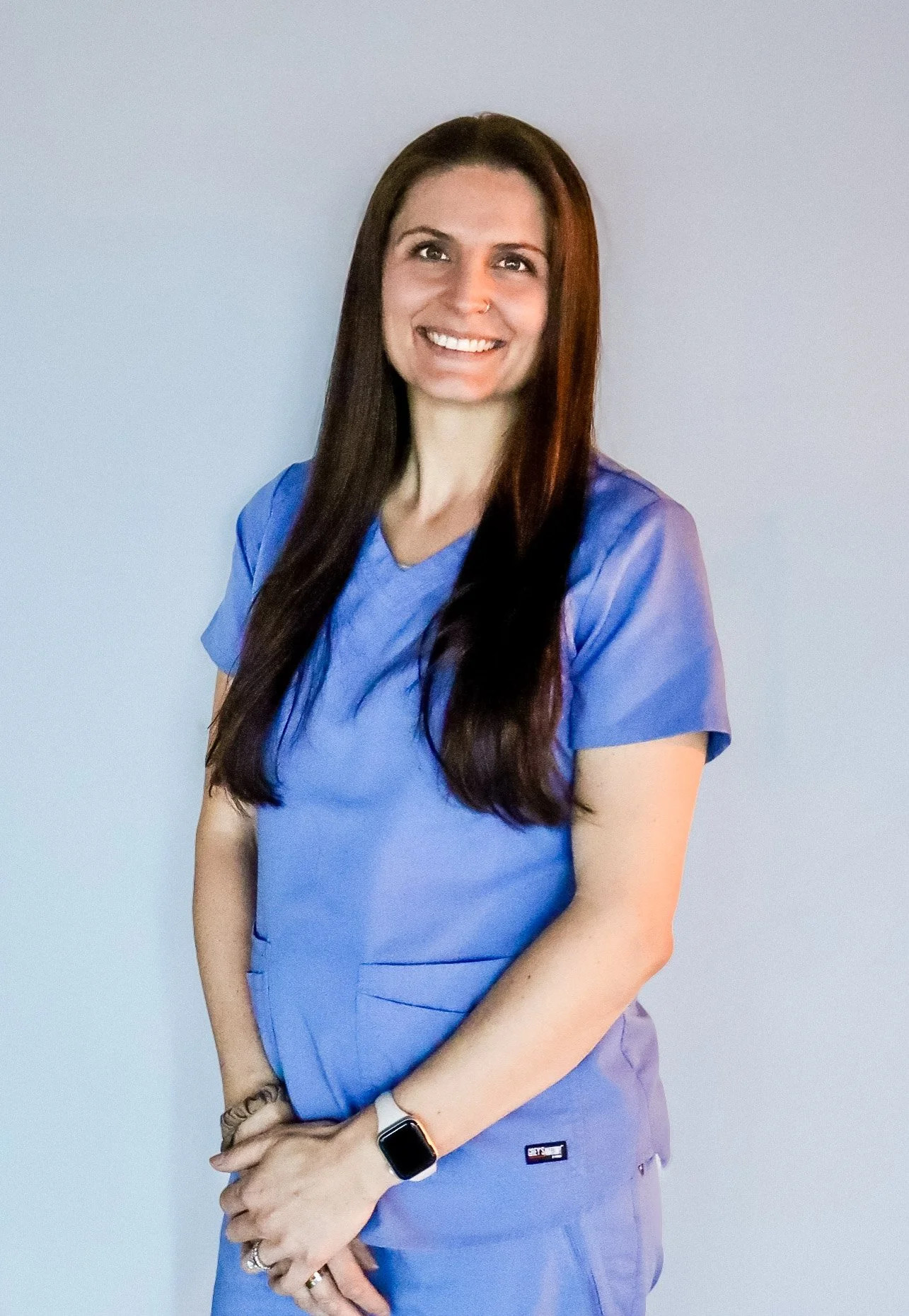 A woman with long dark hair wearing blue medical scrubs, smiling, standing against a light gray background.