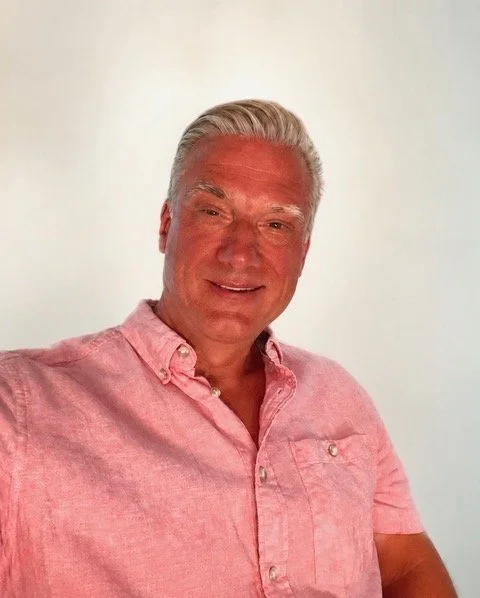 Portret van Dennis Verheugd, die een roze overhemd draagt, glimlachend voor een witte achtergrond die uitvaart muziek verzorgt.