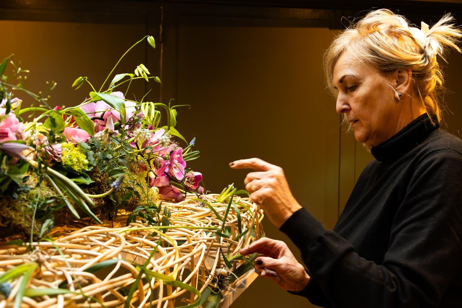 Een vrouw die een rouwboeket verzorgt op een uitvaartkist met kleurrijke bloemen, waarschijnlijk in een binnenomgeving.