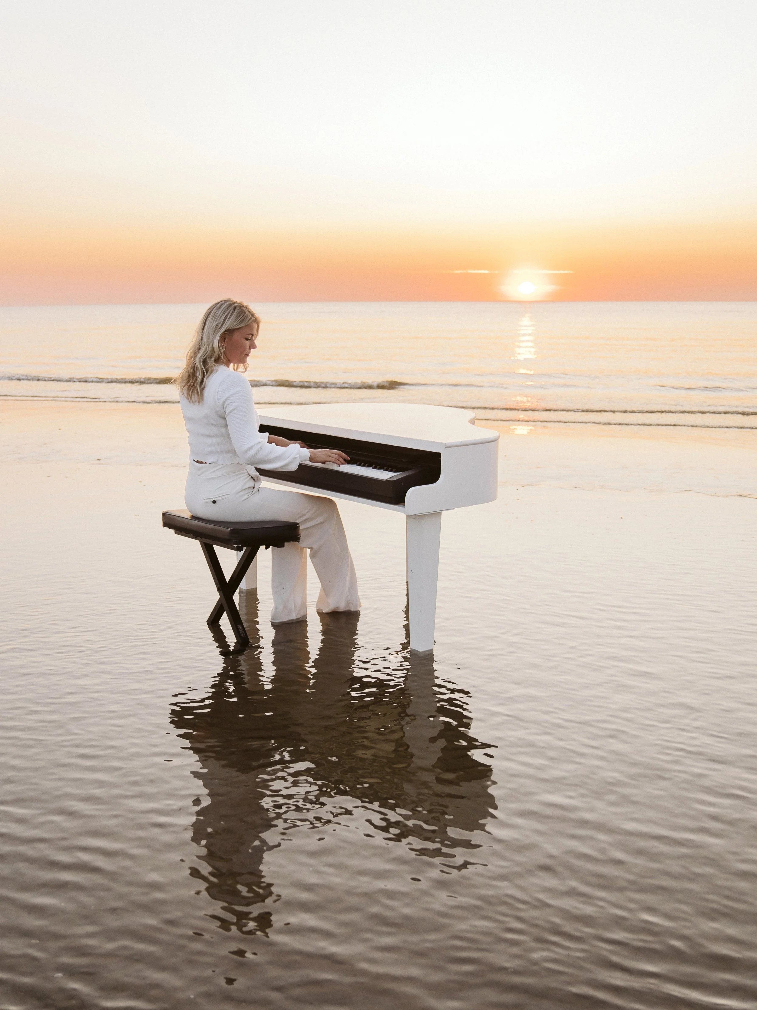 Livemuziek voor bij een uitvaart door vrouw die piano speelt in water tijdens zonsondergang op strand.