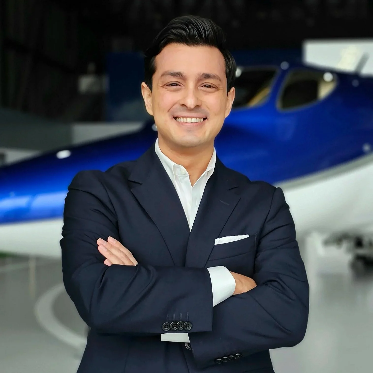 A man in a dark suit with crossed arms smiling in front of a blue airplane.