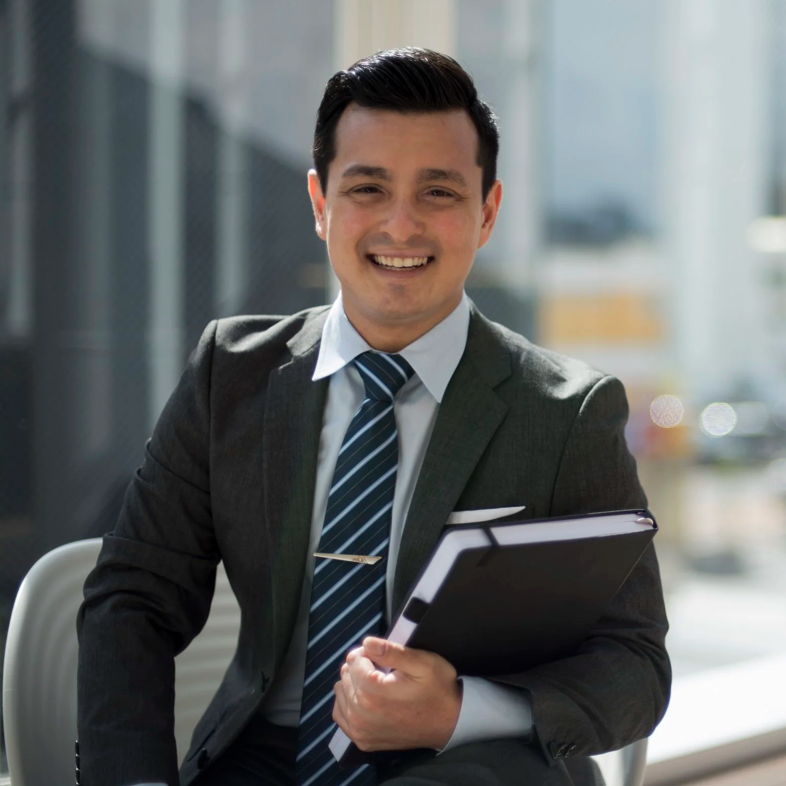 Young man in business attire smiling, holding a portfolio, in a modern office building.