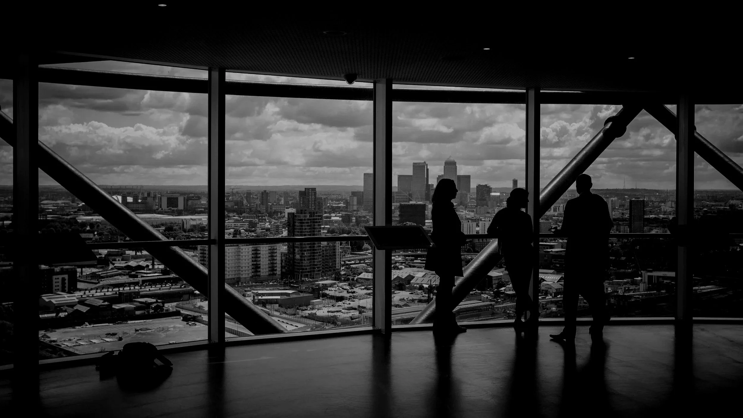 Silhouetted people talking inside a modern skyscraper with large windows, overlooking a city skyline with tall buildings and cloudy sky.
