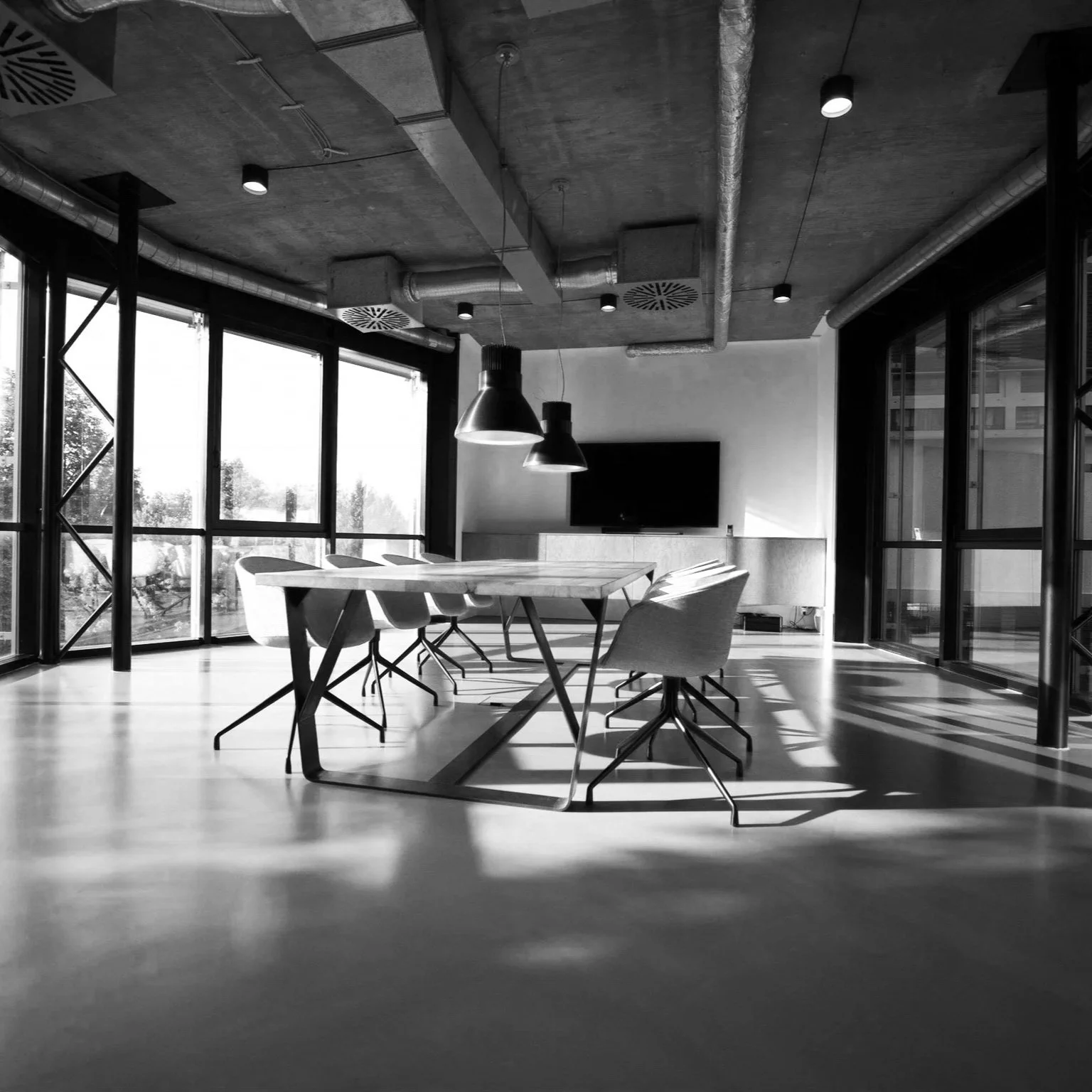 Modern conference room with a large wooden table, six chairs, and a wall-mounted TV, with floor-to-ceiling windows allowing natural light and city view, black-and-white photo.